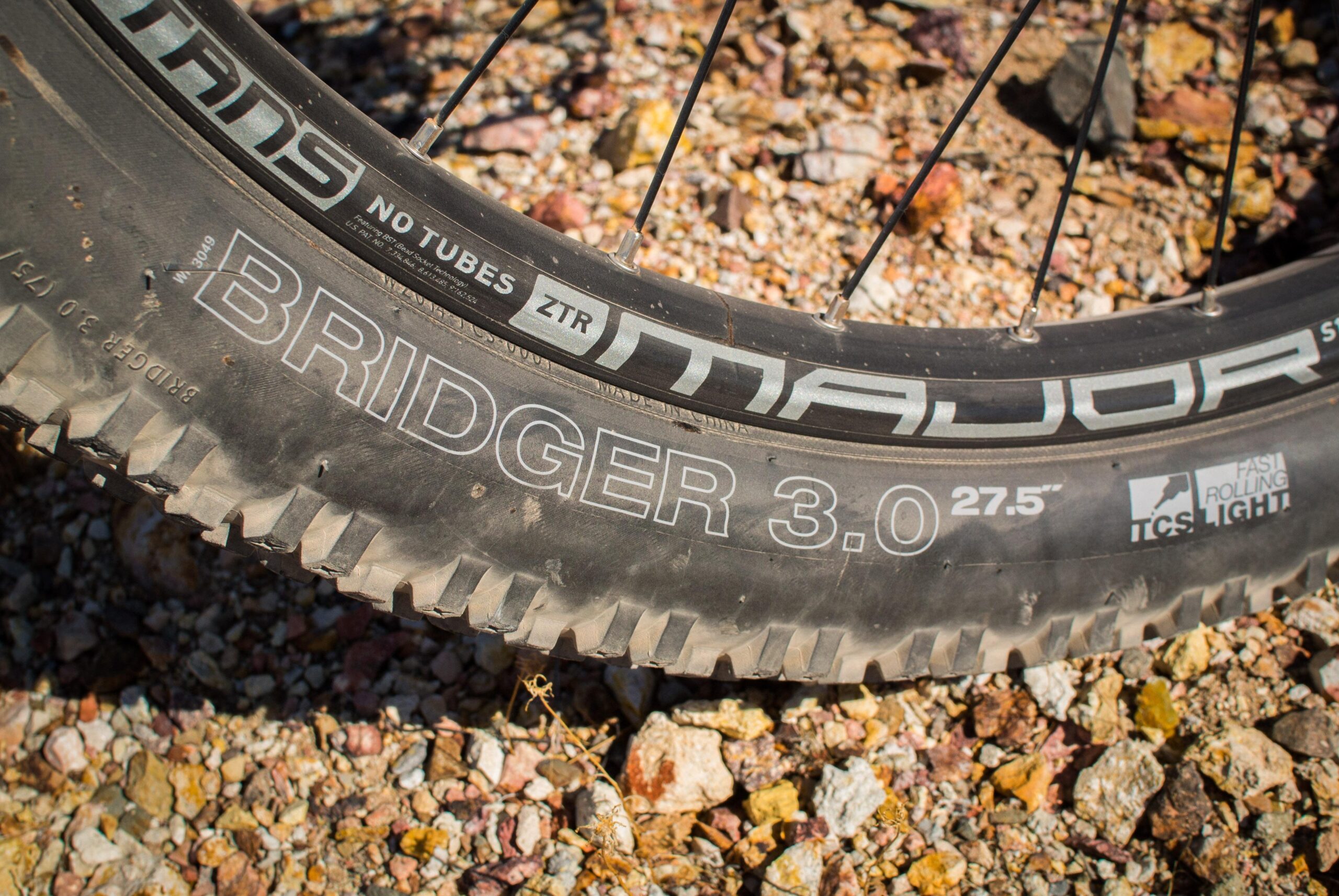 Marin B-17 3: Close-up of a mountain bike tire labeled "Bridger 3.0" on a gravel surface, featuring "No Tubes ZTR" and "27.5" markings, with visible tread pattern and a rugged terrain backdrop.