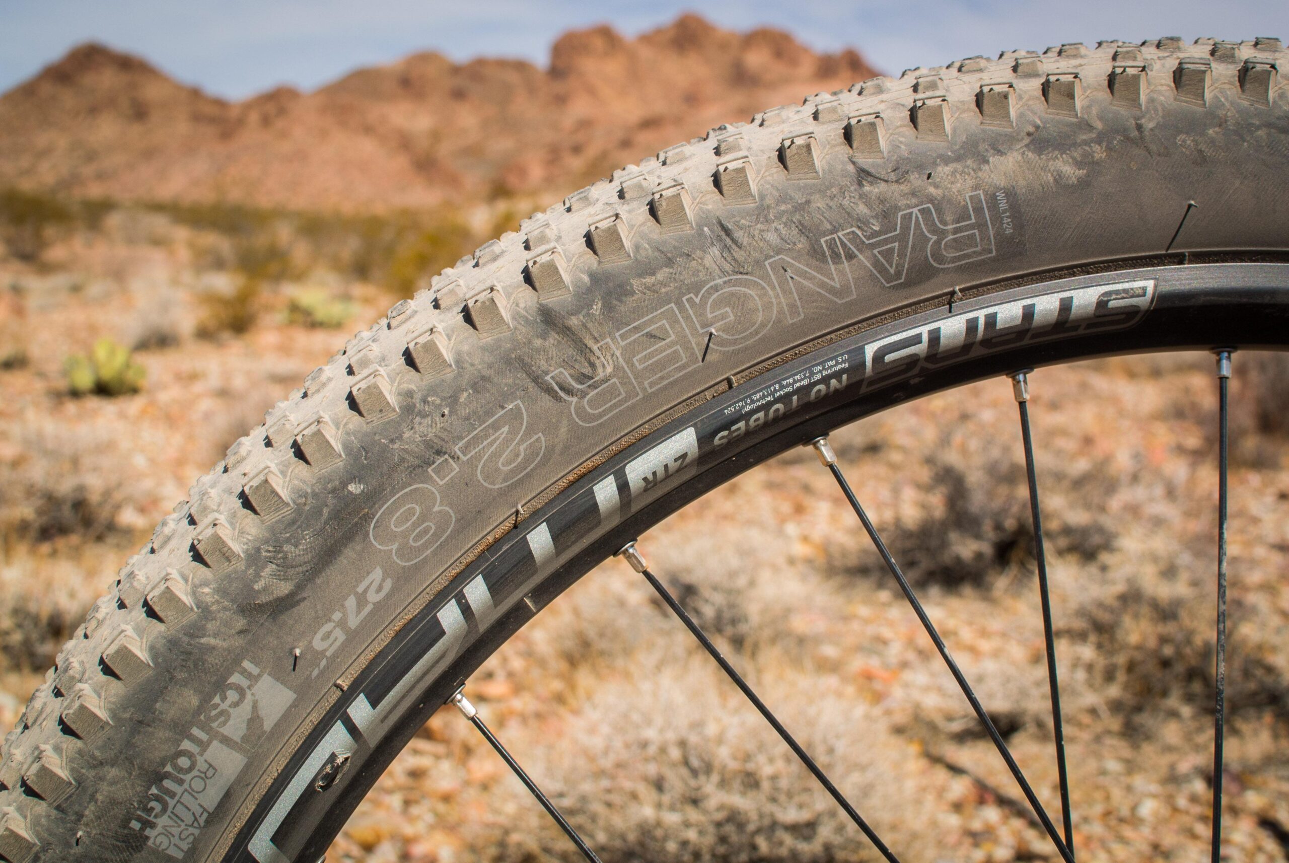 Marin B-17 3: A close-up view of a mountain bike tire on a rocky terrain, highlighting its textured tread and sidewall markings, with blurred mountains in the background.