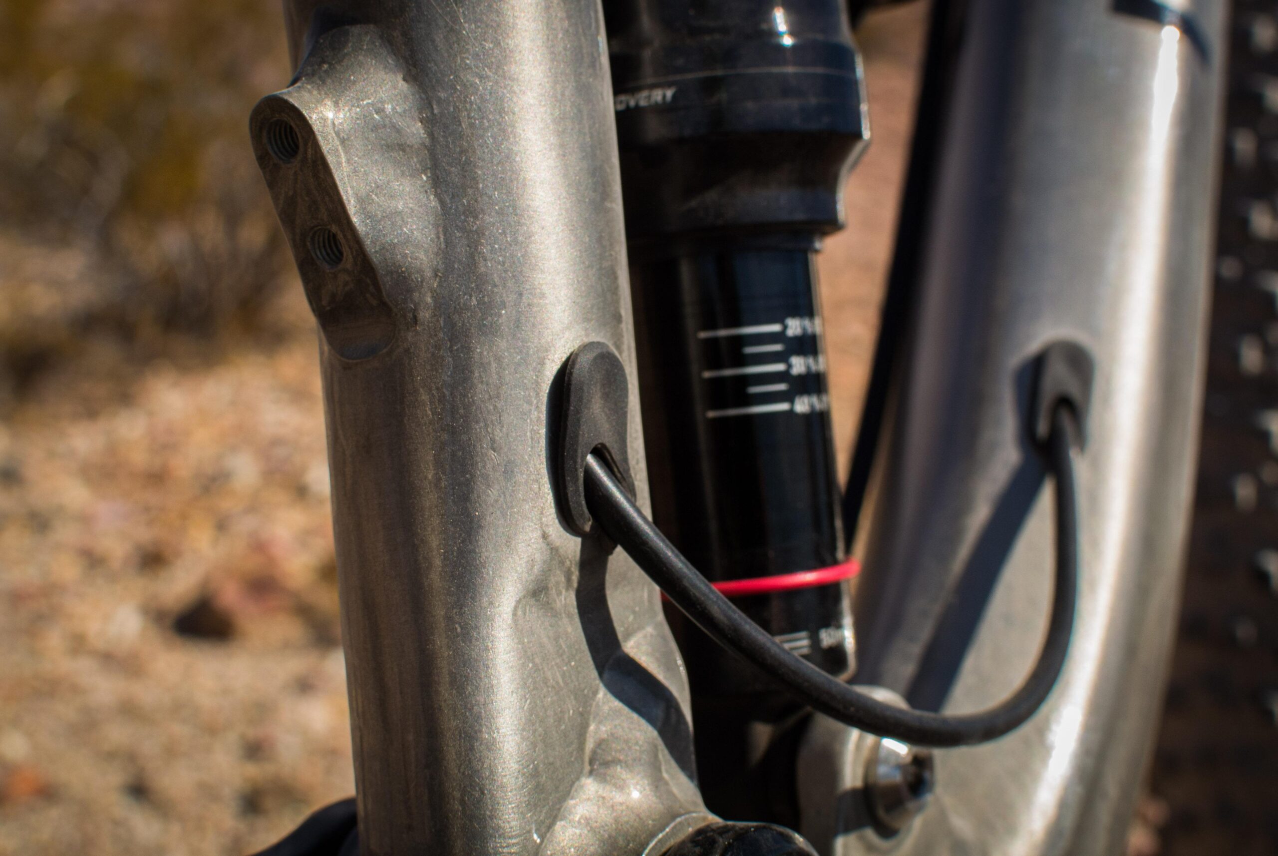 Marin B-17 3: Close-up view of a mountain bike's frame, showcasing the suspension system. The image highlights a black cable routed through a rubber guide, attached to a metallic frame with a brushed finish. In the background, the blurred outline of natural terrain is visible.