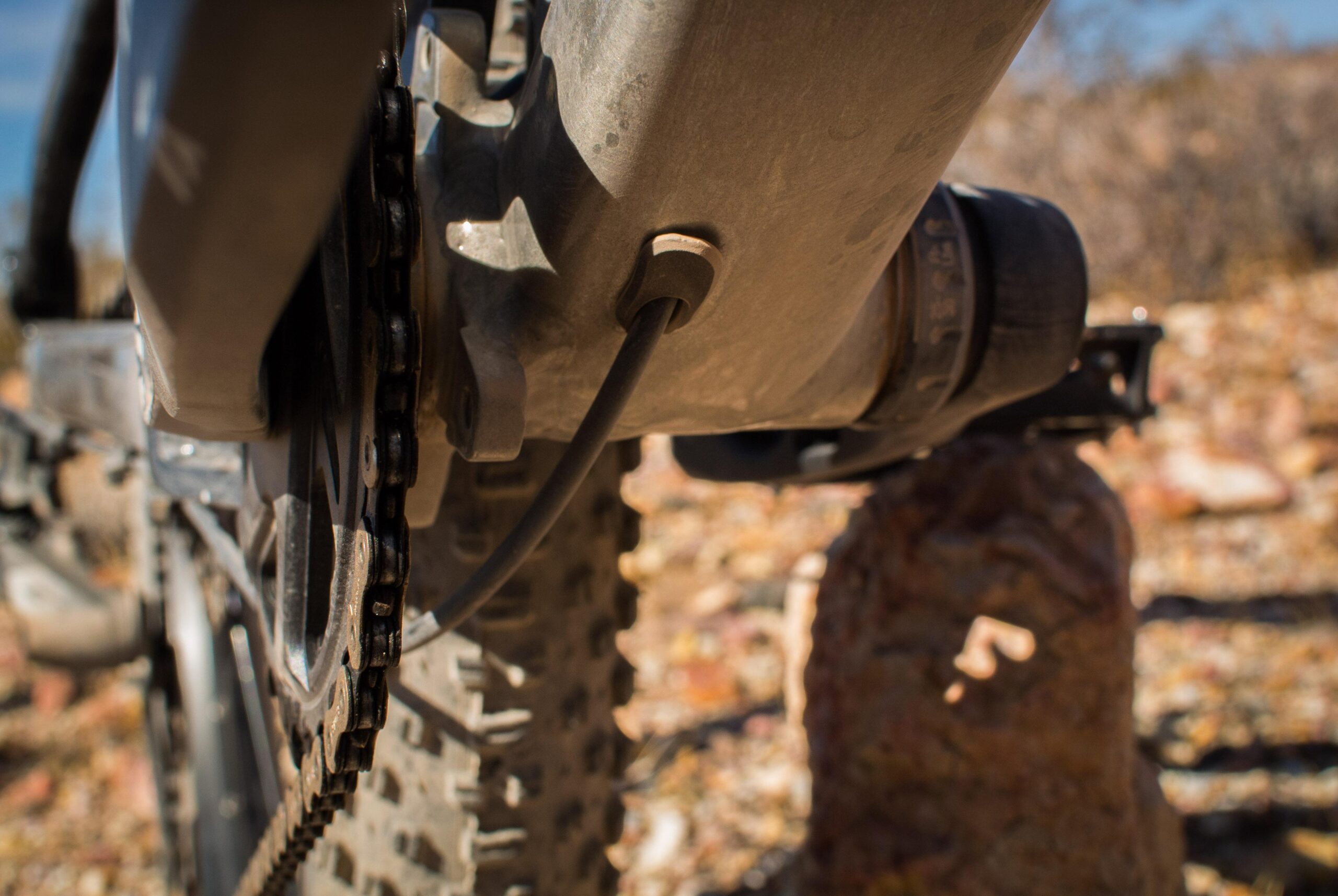 Marin B-17 3: Close-up view of a bicycle's drivetrain, featuring the chain, gear, and a portion of the bike frame against a rugged outdoor background. The image highlights details such as the bike's chain and components, with a dusty, natural setting visible in the background.