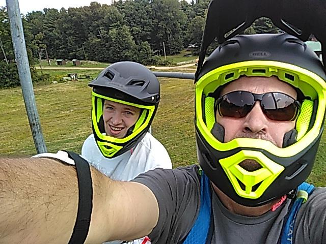 Two individuals wearing bright yellow and black helmets sit on a ski lift, smiling for a selfie. They are surrounded by a green landscape with trees in the background. The atmosphere appears cheerful and adventurous. West Mountain mountain bike trail.