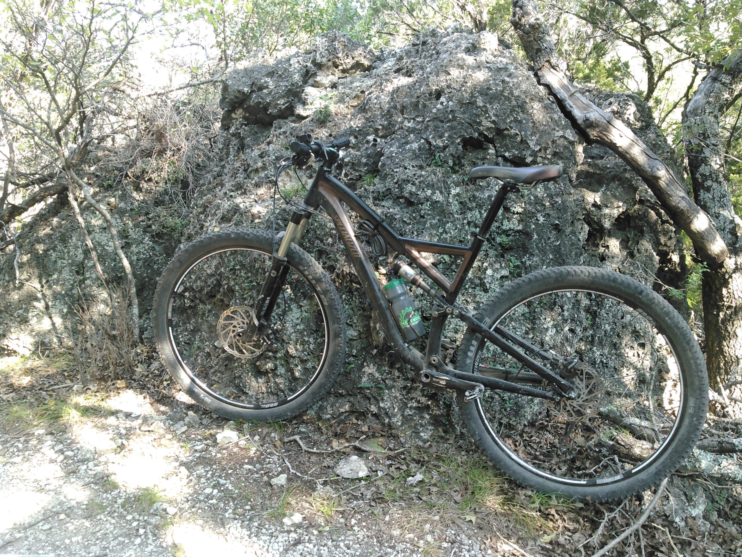 A mountain bike leaning against a large rock, surrounded by trees and greenery on a dirt trail. The bike has a dark frame and is equipped with wide tires, while a water bottle is attached to the frame. Sunlight filters through the leaves, casting dappled shadows on the ground. Dana Peak mountain bike trail.