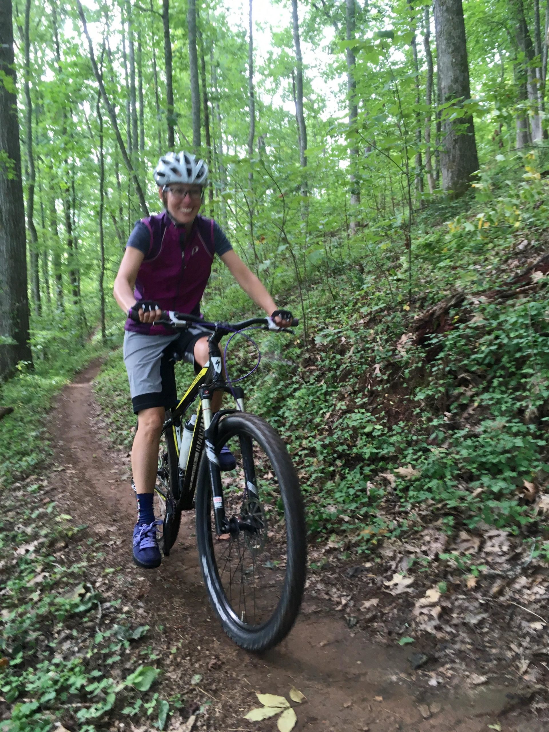 A cyclist wearing a helmet and a purple vest rides a mountain bike along a dirt trail in a lush green forest. The cyclist is smiling and appears to be enjoying the ride. Surrounding trees and foliage create a vibrant natural setting. Jack Rabbit Trails mountain bike trail.