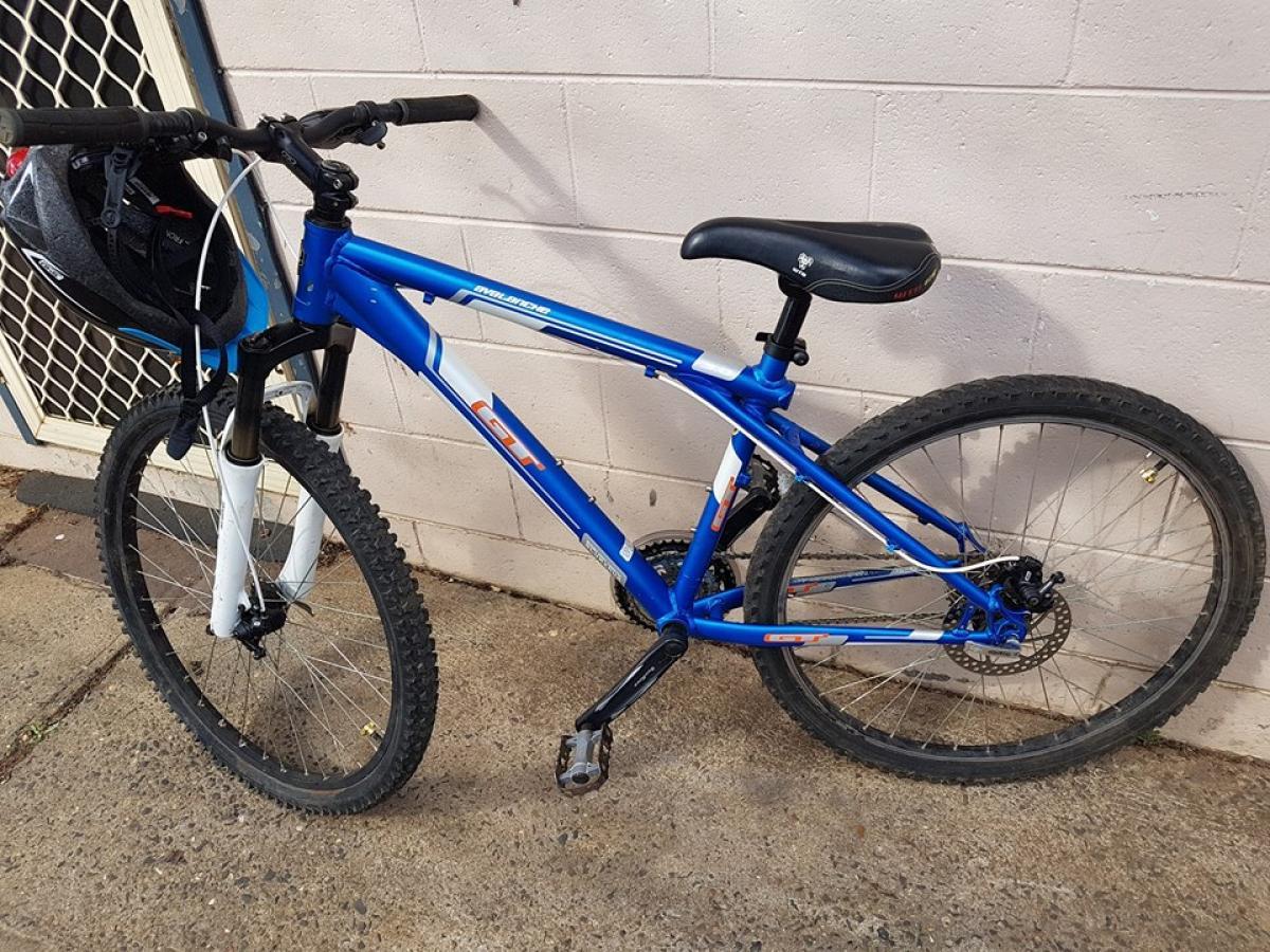 GT Avalanche: A blue mountain bike with a black seat, located against a light-colored wall. The bike features thick tires and front suspension, with a helmet hanging from the handlebars. The background includes a textured surface and an entrance with a grille.