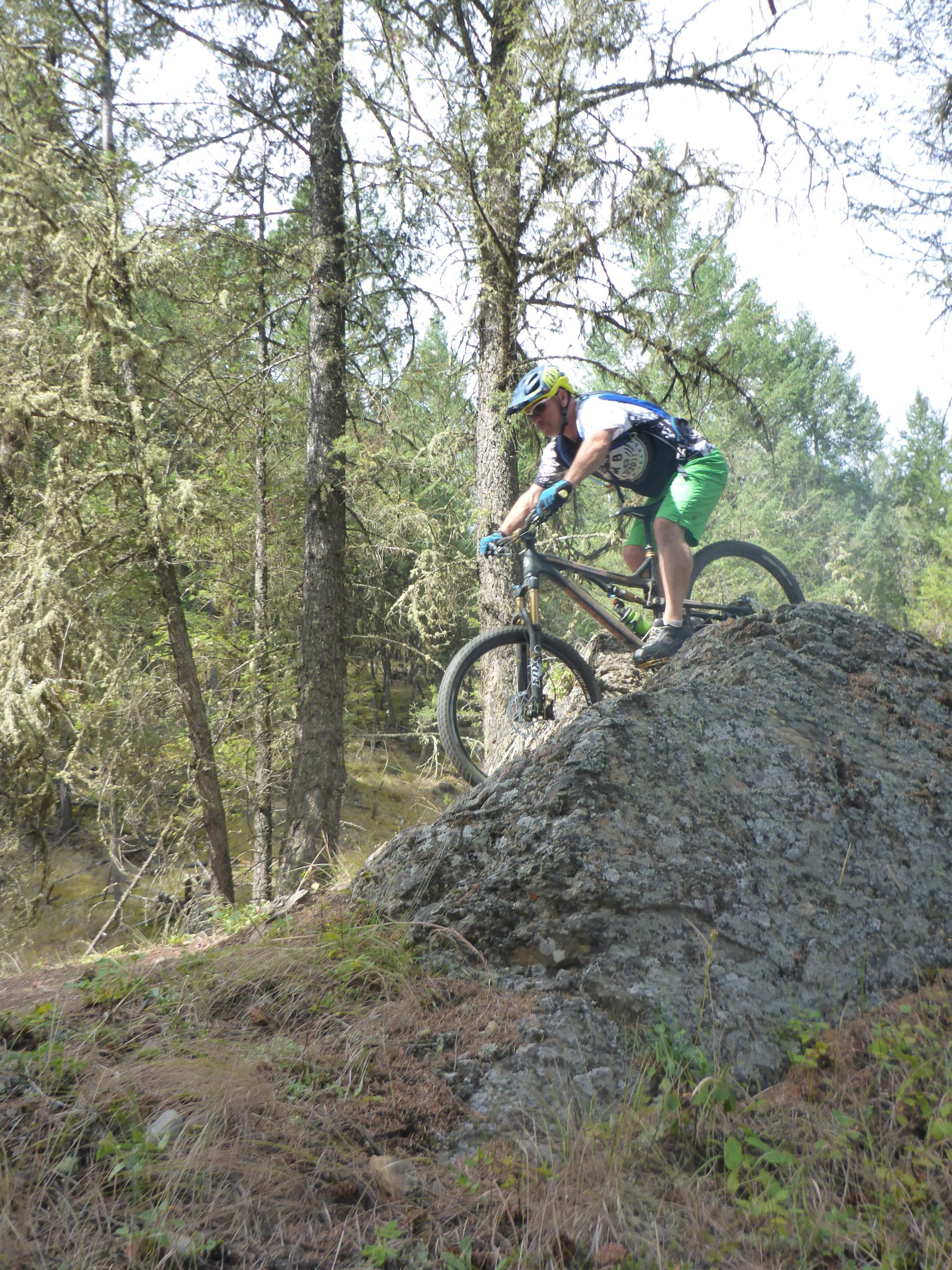 A mountain biker in a helmet and casual gear expertly navigates rocky terrain in a forested area, showcasing a thrilling moment as they ride over a large boulder surrounded by trees and greenery. Lake Lillian mountain bike trail.