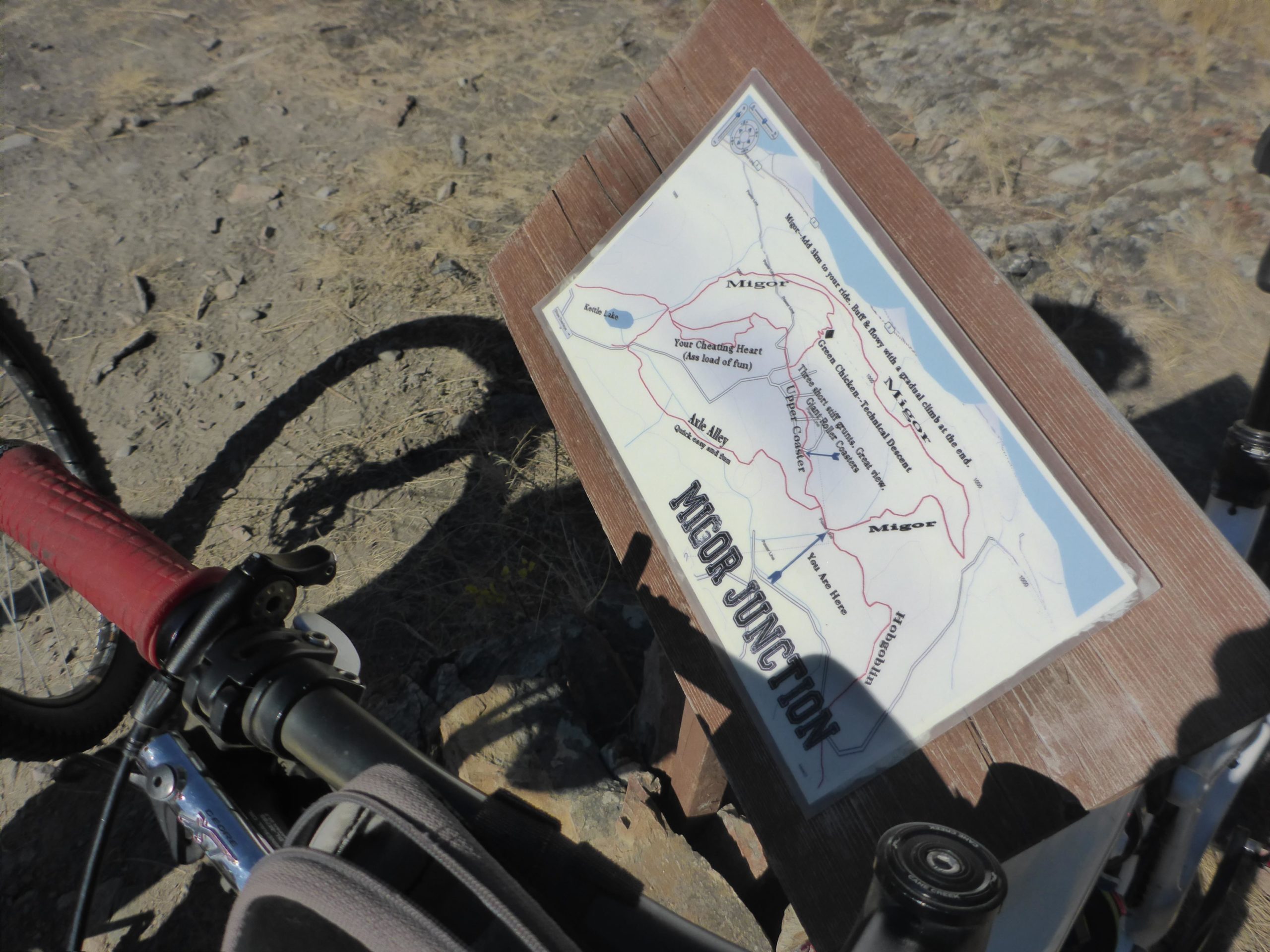A close-up view of a mountain bike positioned on a dirt path next to a trail map sign titled "Major Junction." The map outlines various biking routes in the area, with red lines indicating the trails. The ground is rocky and there are patches of dry grass around. Cranbrook Community Forest - South mountain bike trail.
