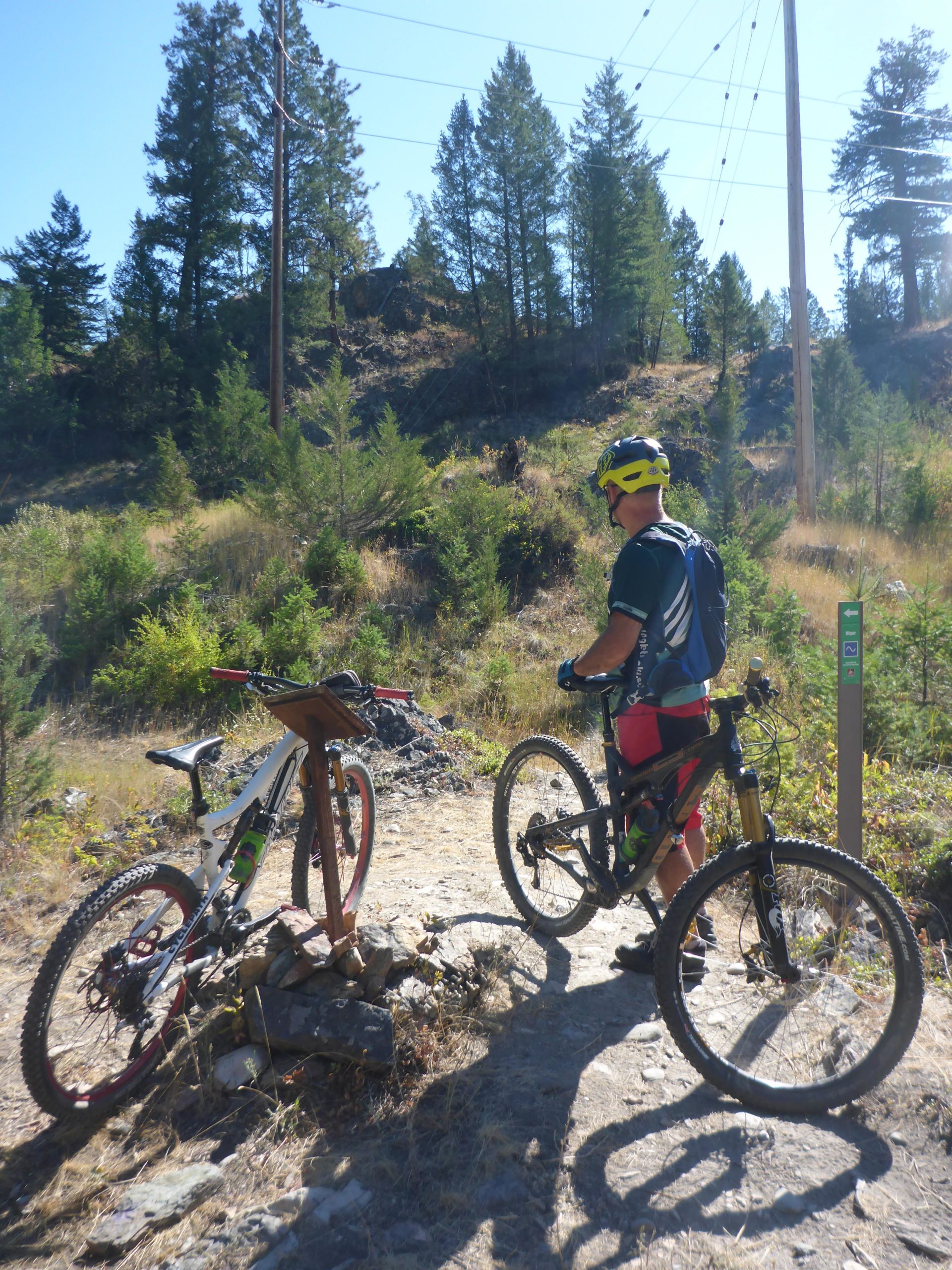 A mountain biker stands on a rocky trail, looking at two bicycles positioned near a trail marker. Surrounding him is a landscape of tall trees and grassy underbrush, with power lines visible in the background under a clear blue sky. Cranbrook Community Forest - South mountain bike trail.