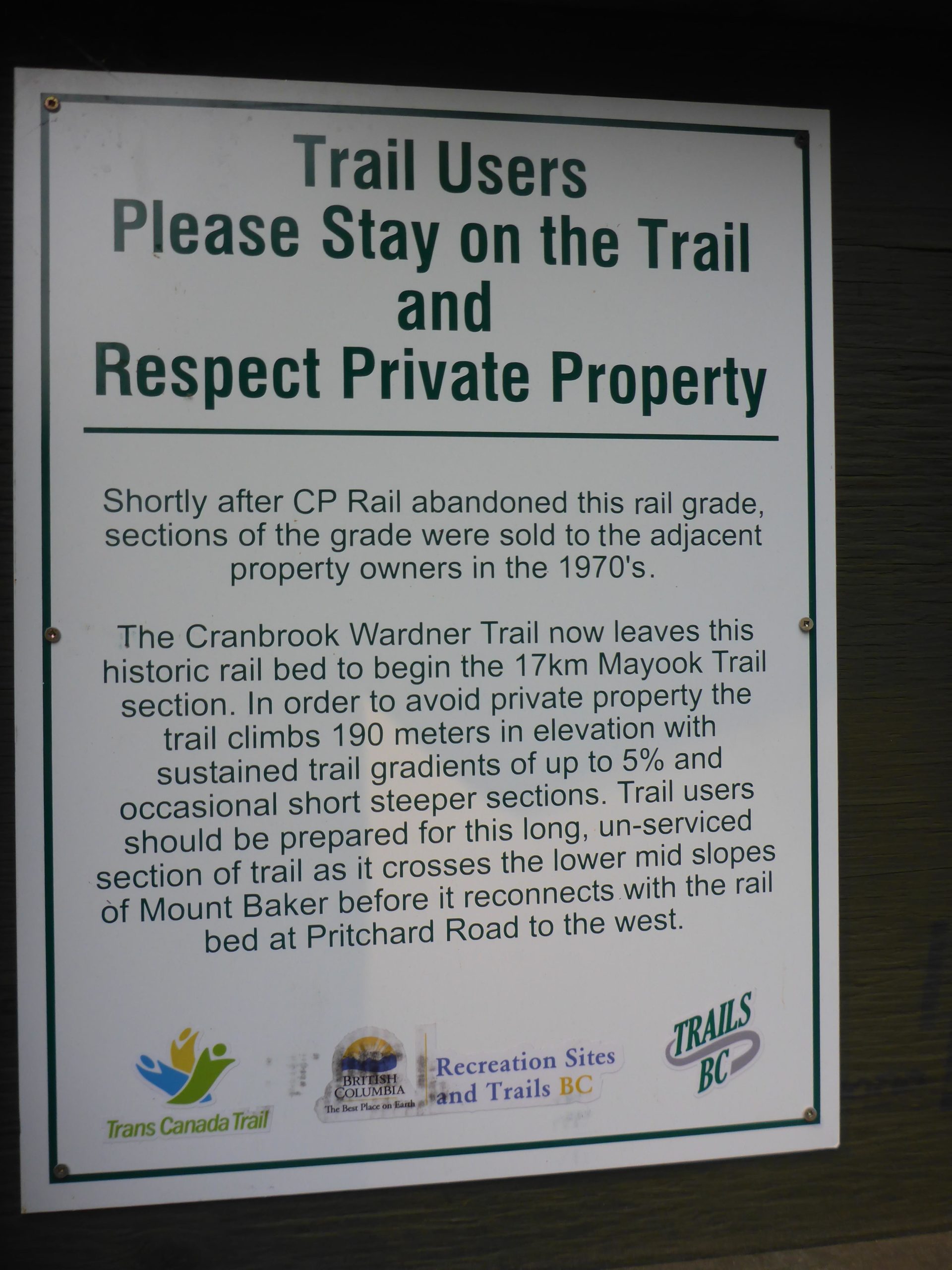 Sign indicating trail usage guidelines, advising users to stay on designated trails and respect private property. It describes the Cranbrook-Wardner Trail and the Mayook Trail section, including elevation details and trail conditions, along with logos from organizations like Trans Canada Trail and Recreation Sites and Trails BC. Mayook Trail mountain bike trail.