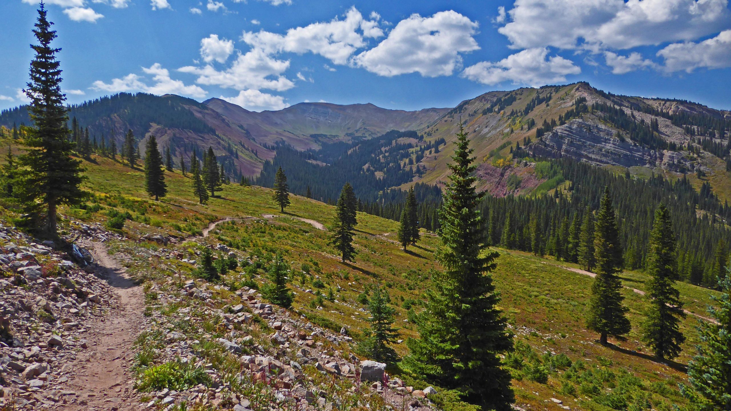 A scenic view of a mountain landscape featuring a winding dirt path surrounded by green meadows and coniferous trees. The backdrop includes towering mountains under a blue sky with fluffy white clouds. The vibrant colors of the foliage and rocky terrain create a tranquil and natural setting. Bike Snowmass Bike Park mountain bike trail.