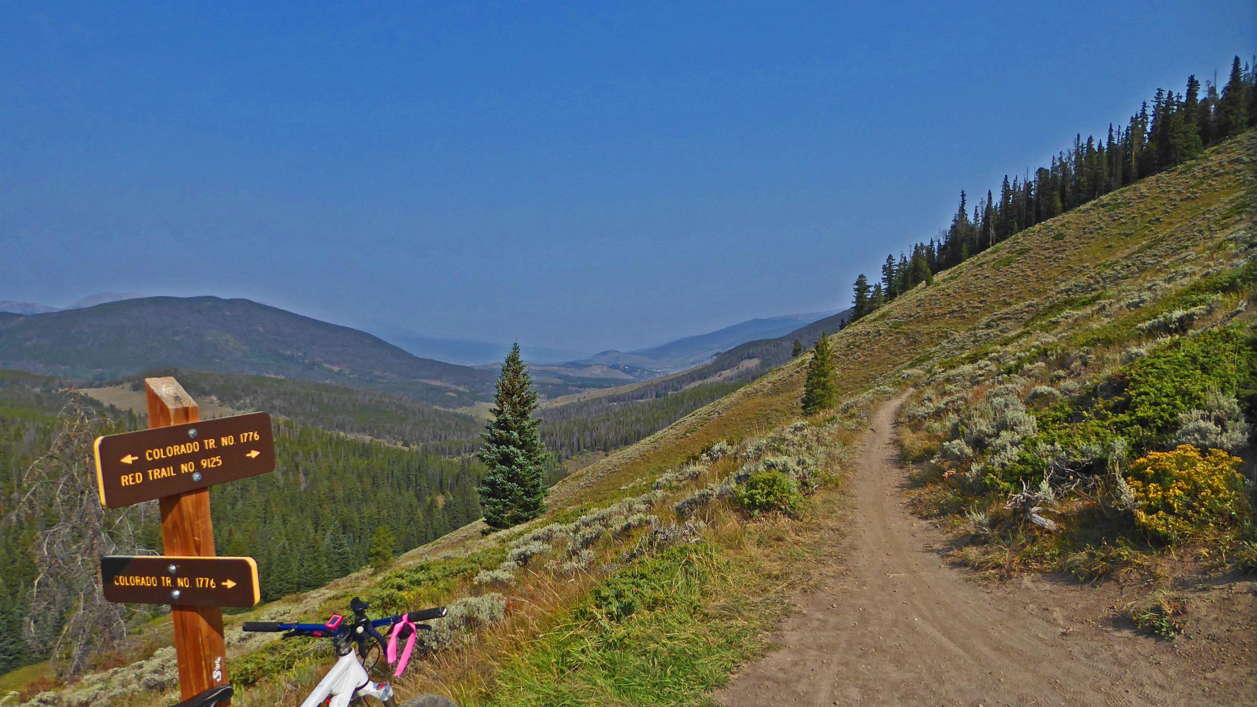 A scenic view of a mountain trail with signage indicating various trail routes. A mountain bike rests near the sign on the left, while a vibrant landscape of rolling hills, trees, and distant mountains stretches into the background under a clear blue sky. The trail winds along the hillside, showcasing the lush greenery of the surrounding area. Red Trail mountain bike trail.