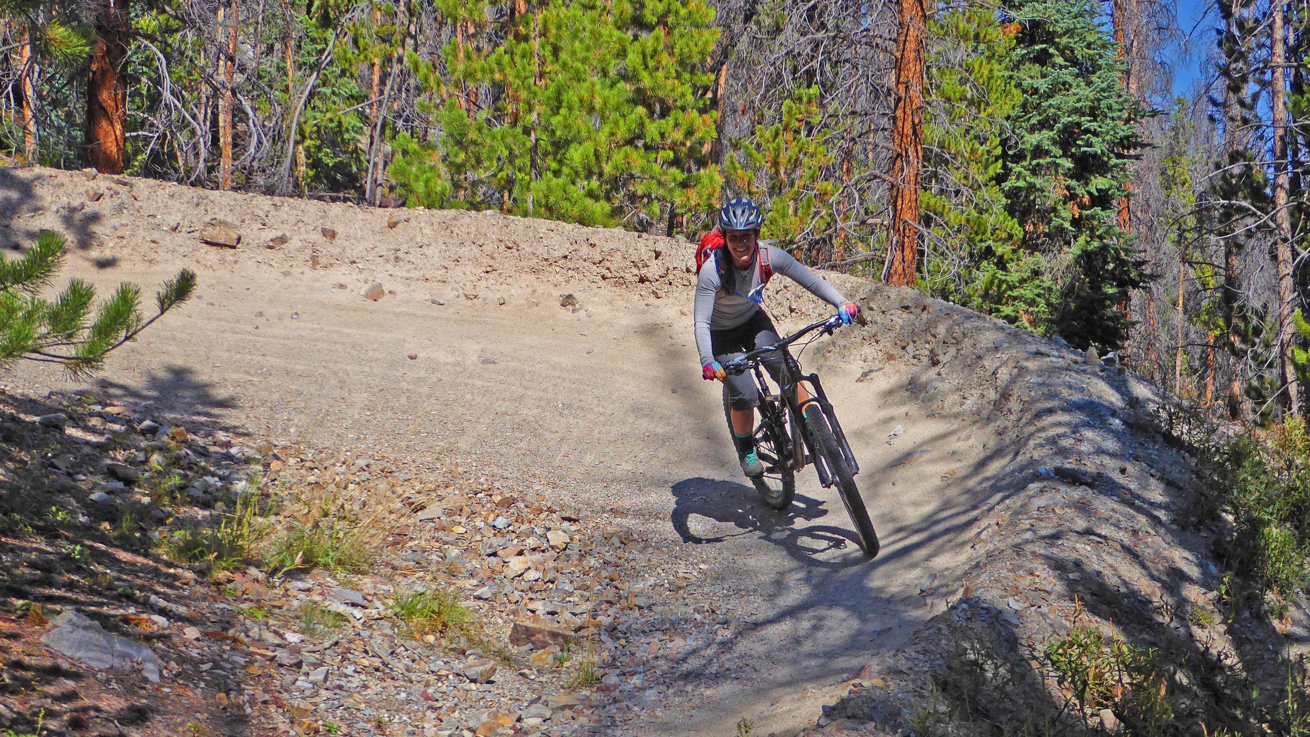 A mountain biker riding along a winding dirt trail surrounded by trees and underbrush, smiling as they navigate the terrain. The trail has a rocky surface, and the sun shines brightly, illuminating the vibrant green of the surrounding foliage. Slalom mountain bike trail.