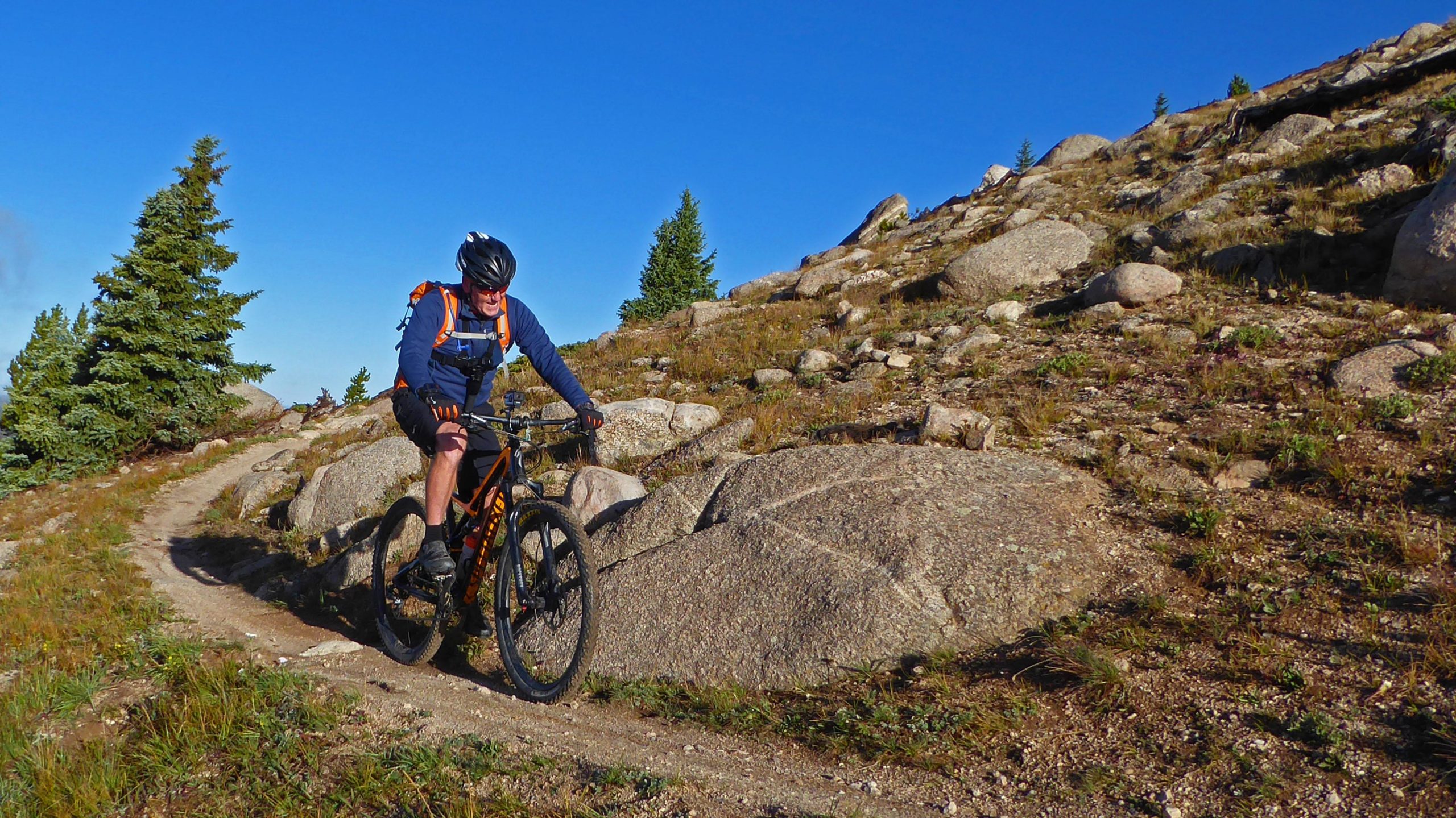 A mountain biker navigating a rocky trail, surrounded by lush greenery and boulders under a clear blue sky. The biker is wearing a helmet and a backpack, focused on riding along the rugged terrain. Monarch Crest Trail mountain bike trail.