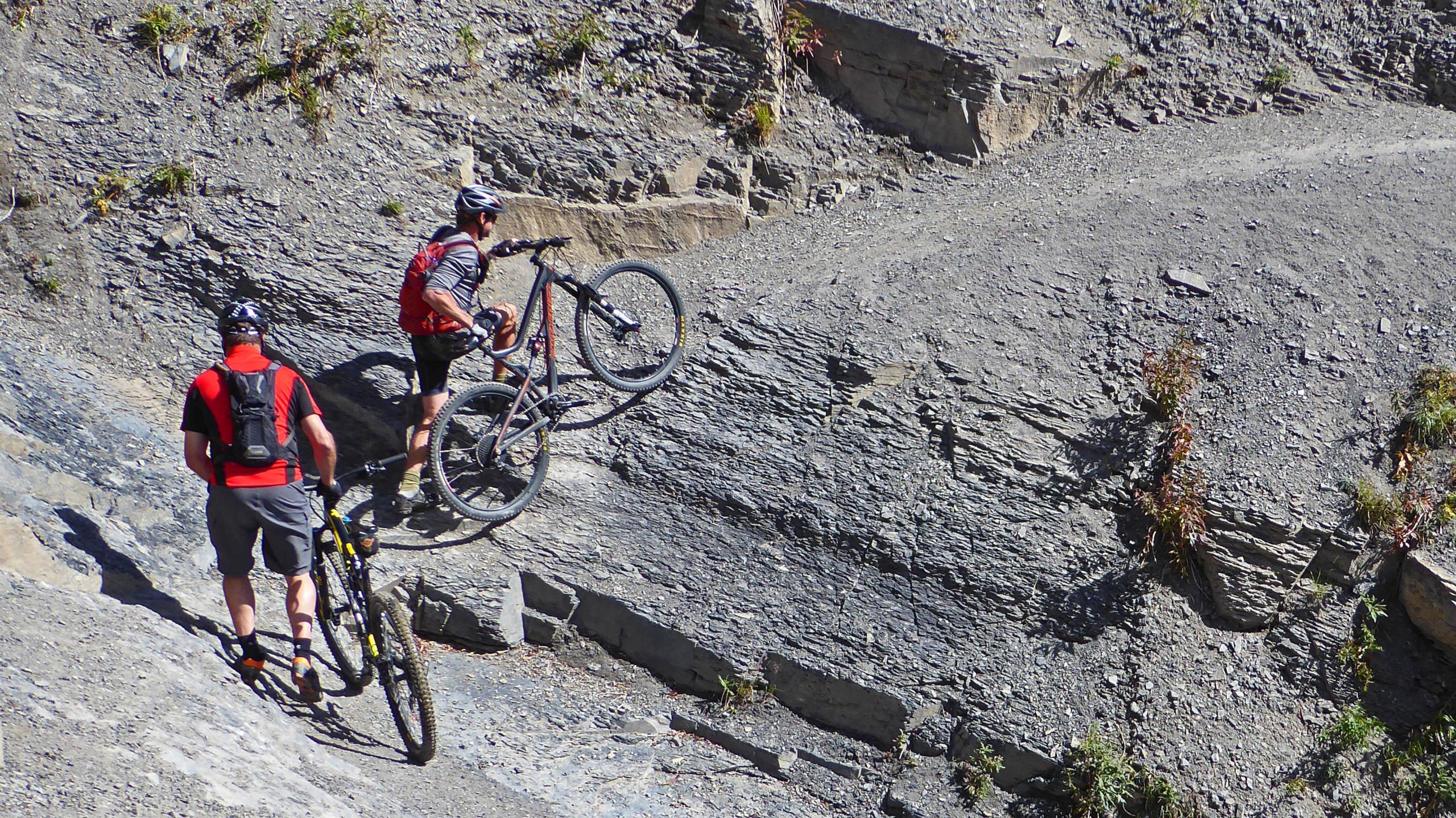 Two mountain bikers navigating a rocky trail, one balancing with their bike on one wheel while the other walks with their bike beside them. The terrain is rugged with loose gravel and sparse vegetation. Trail 401 mountain bike trail.