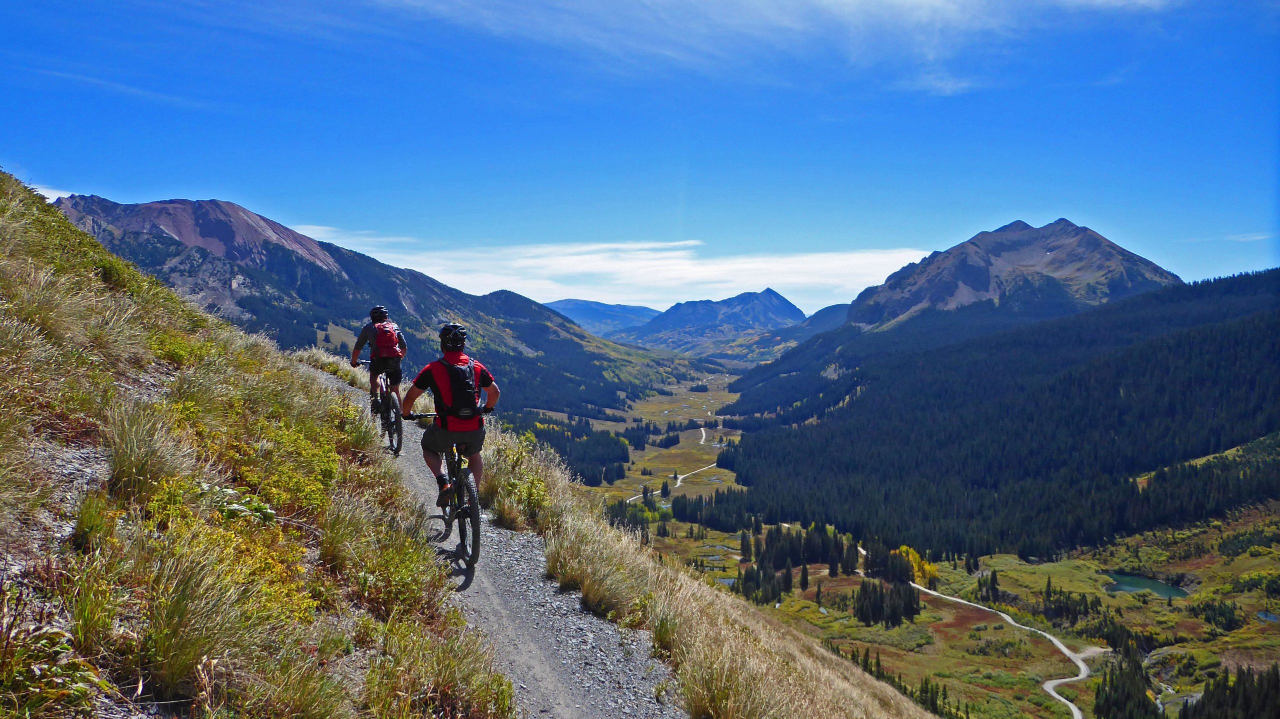Two mountain bikers riding along a dirt trail high in the mountains, surrounded by lush greenery and panoramic views of valleys and peaks under a clear blue sky. Trail 401 mountain bike trail.