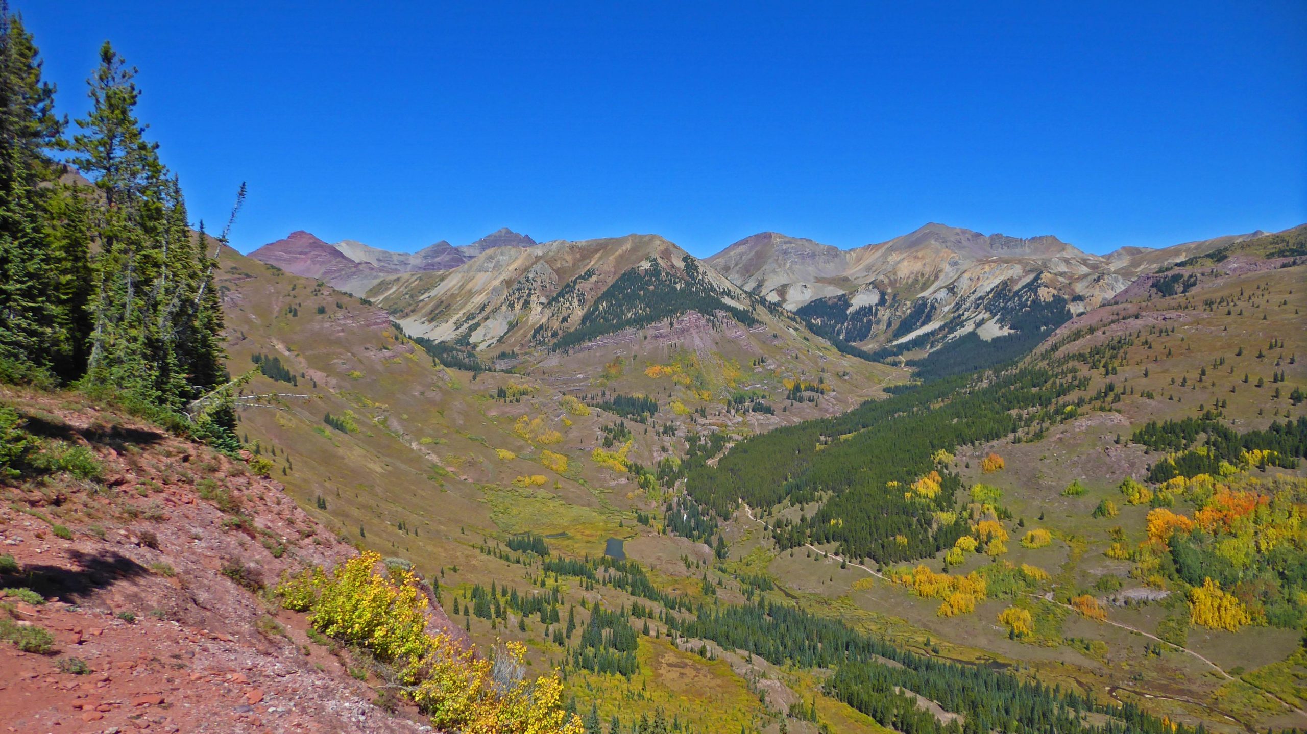 A panoramic view of a mountainous landscape with a clear blue sky. The scene features rolling hills and peaks, some with rocky outcrops and patches of trees, including evergreens and areas of colorful autumn foliage. The valley below includes a lush forest and open fields, showcasing a blend of green and golden hues. Teocalli Ridge mountain bike trail.