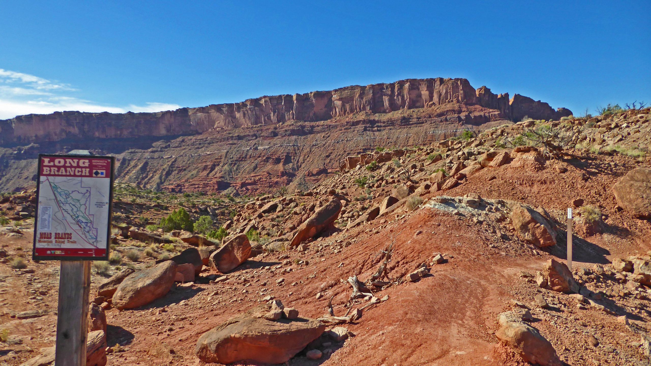 A rugged landscape featuring red rocky terrain under a blue sky, with a prominent cliff face in the background. A trail sign labeled "Long Branch" is visible in the foreground, providing information about the trails in the area. The scene conveys a sense of natural beauty and adventure. Long Branch mountain bike trail.