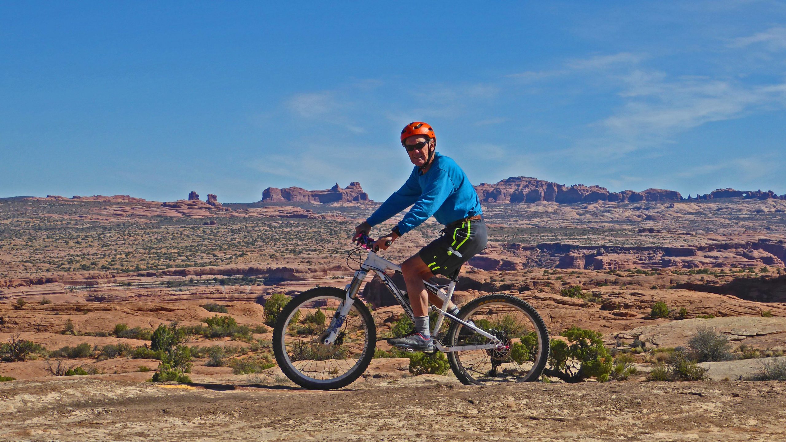 A person riding a mountain bike on rocky terrain with a desert landscape in the background, featuring rugged cliffs and a clear blue sky. The rider is wearing a blue long-sleeve shirt, black shorts with bright green detailing, an orange helmet, and sunglasses. Rockin' A mountain bike trail.