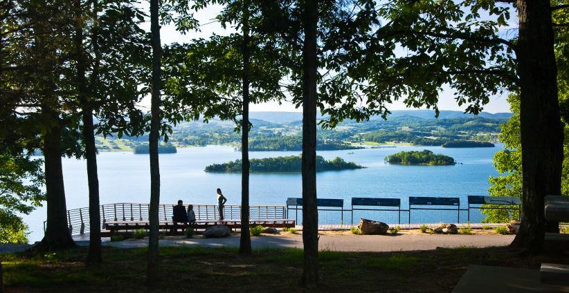 A scenic view of a lake surrounded by green hills, with two people sitting on a bench in the foreground. The area is framed by trees, and a small island is visible in the water. The landscape captures a peaceful, natural setting with clear skies and distant mountains. Panther Creek State Park mountain bike trail.