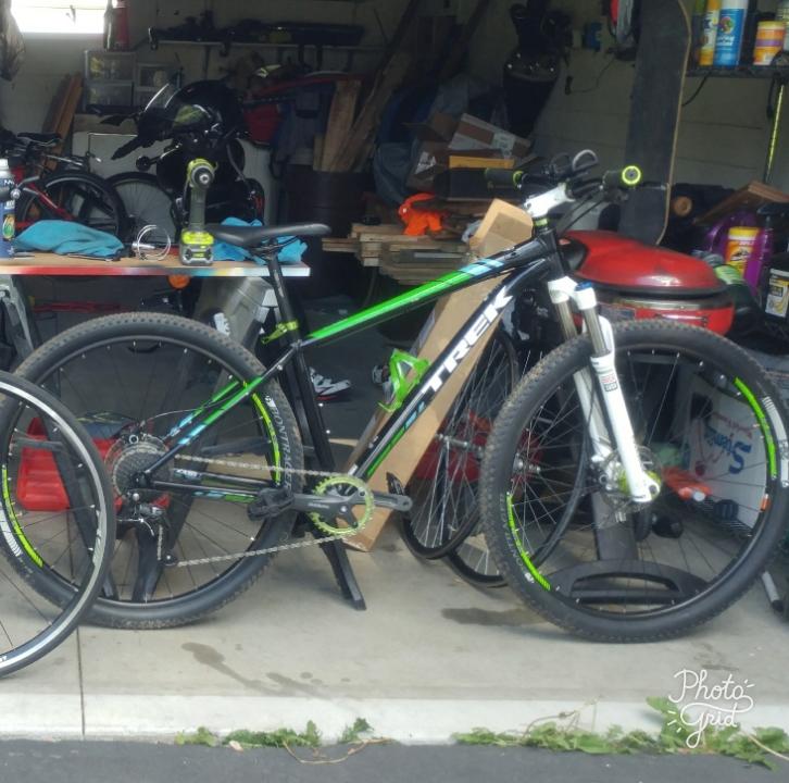 Trek Xcaliber 7: A garage scene featuring two bicycles, with one prominently displayed in the foreground. The bike has a black frame with green accents and is equipped with knobby tires. Various tools and items are visible in the background, along with wooden planks and a red tool box. The setting appears to be an organized workspace for bike maintenance or repair.