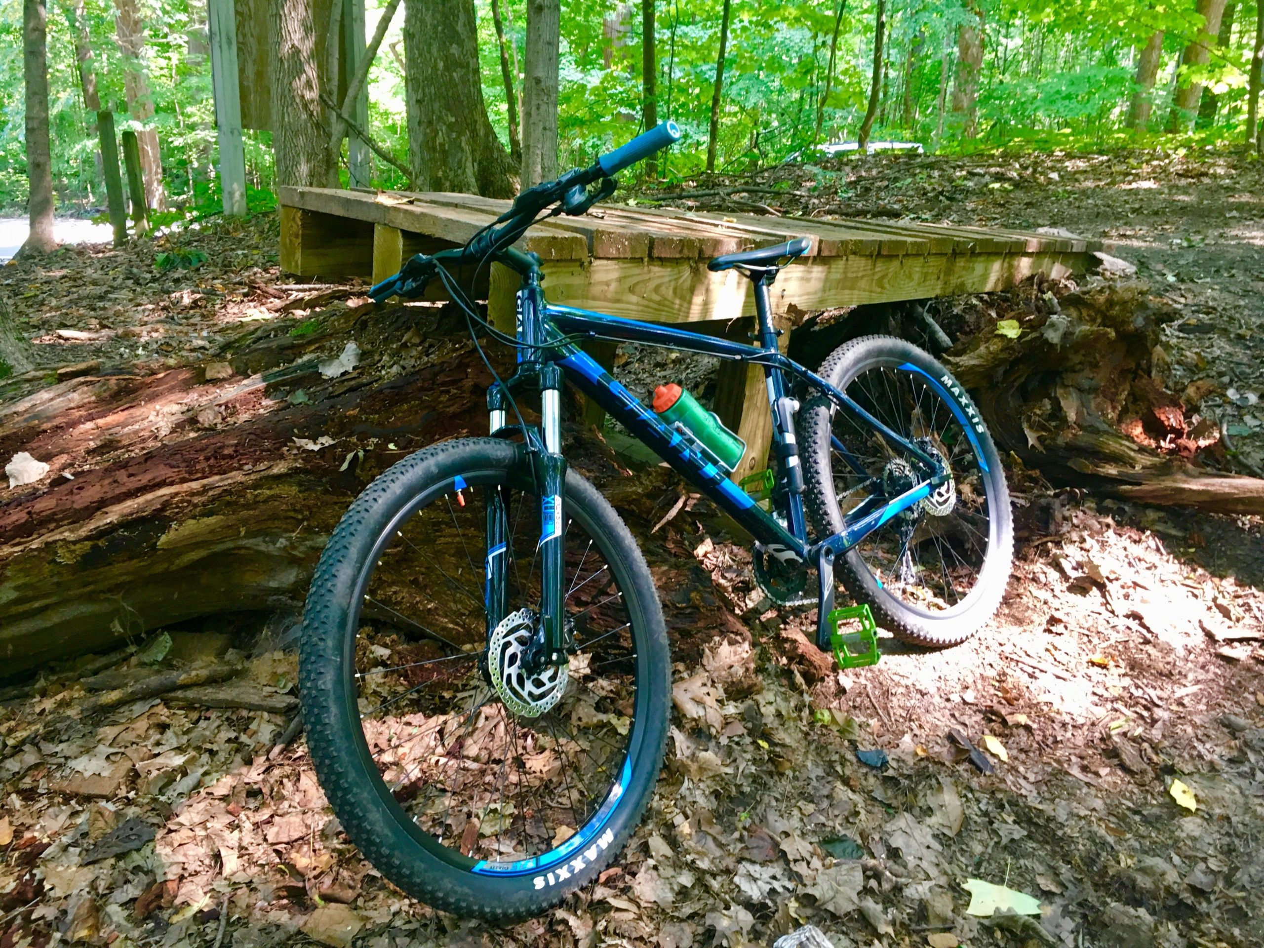 A blue mountain bike resting on a fallen log in a forested area, with a wooden bridge visible in the background. The ground is covered in fallen leaves, and sunlight filters through the trees, creating a peaceful outdoor setting. A water bottle is attached to the bike frame. Rum Village Pathway Mountain Biking Trail mountain bike trail.