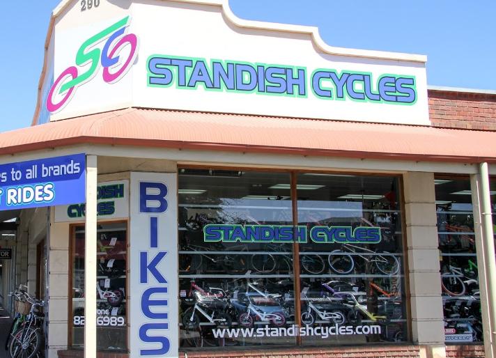 Storefront of Standish Cycles featuring a large sign with the store name, "STANDISH CYCLES," and the word "BIKES." The windows show a variety of bicycles on display. A blue sign nearby indicates services for all brands and offers rides. The building is well-lit with clear skies in the background.