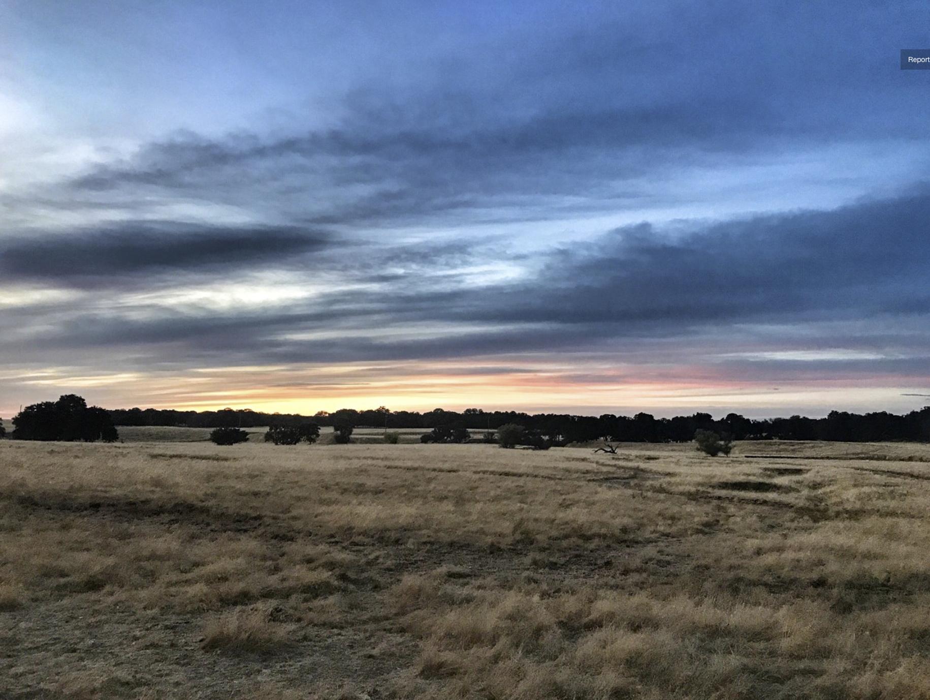 A scenic view of a field at dusk, featuring tall, golden grass and scattered trees silhouetted against a colorful sky with clouds in shades of blue, pink, and gray. The horizon glows softly as the sun sets, creating a peaceful and tranquil atmosphere. Deer Creek Hills: Southeast Loop mountain bike trail.