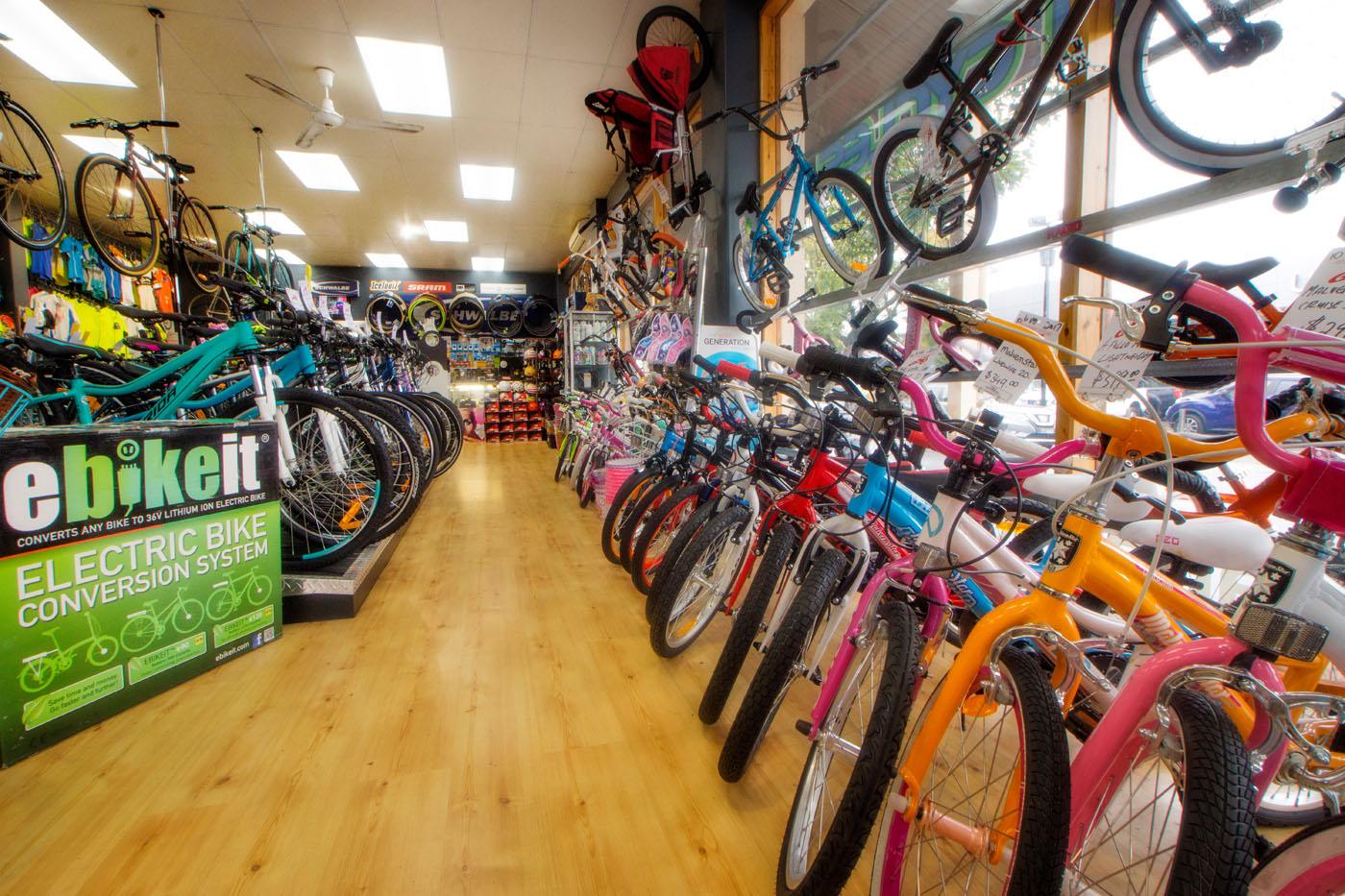 A brightly lit bike shop interior featuring a variety of bicycles displayed along both sides of the aisle. Colorful bikes in shades of pink, blue, and orange are prominent, with some suspended from the ceiling. A sign advertising an electric bike conversion system is visible in the foreground, along with biking accessories showcased on the walls. The wooden floor adds a warm touch to the vibrant environment.