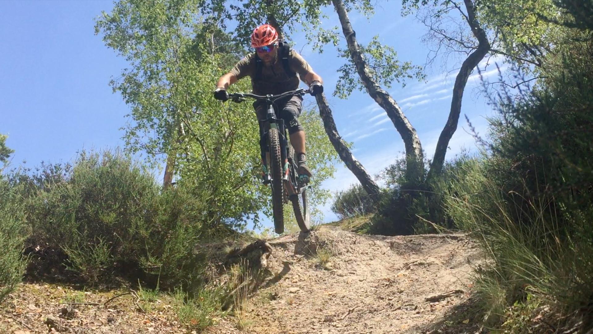 A mountain biker in a red helmet is captured mid-air while jumping over a dirt path surrounded by greenery and trees. The blue sky and sunlit foliage create a bright, energetic atmosphere. Montmorency - XC loop mountain bike trail.