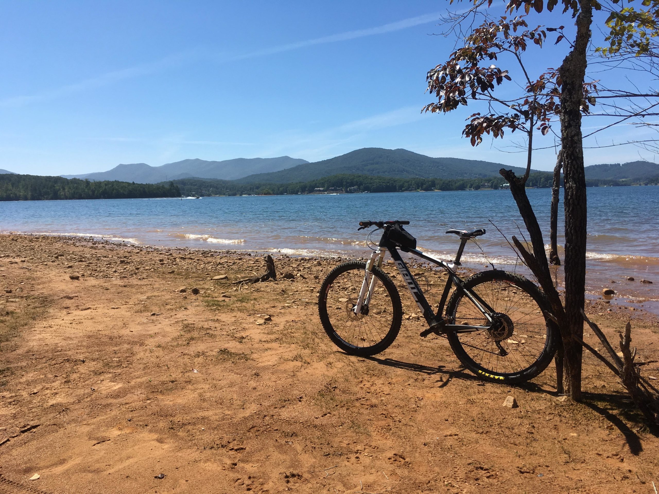 A mountain bike parked on the sandy shore of a lake, with mountains in the background under a clear blue sky. The scene captures a peaceful outdoor setting, ideal for biking and enjoying nature. Jack Rabbit Trails mountain bike trail.