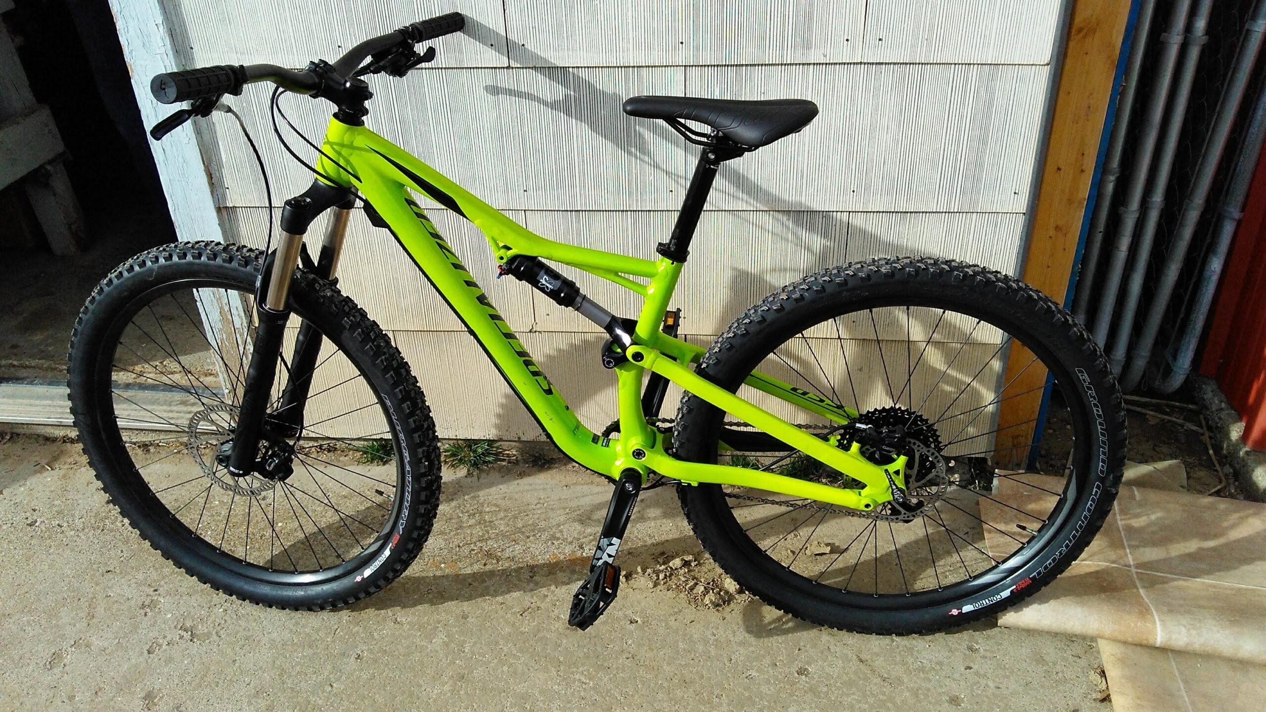Specialized Camber: A vibrant green mountain bike with thick, knobby tires, a black seat, and front suspension forks, positioned against a wooden wall. The bike features a modern frame design and a visibly robust construction, suitable for off-road riding.