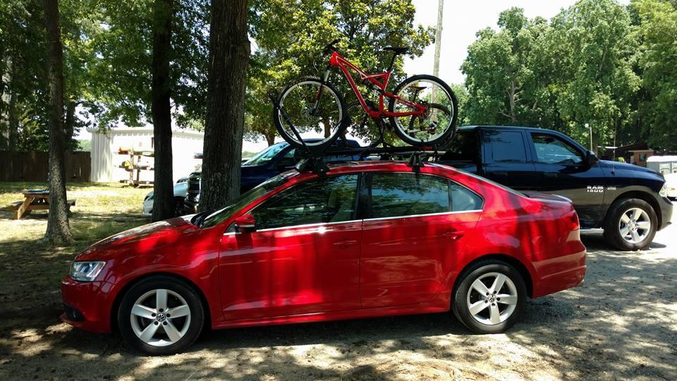 Specialized Camber Comp 29: A red sedan parked under trees, featuring a mountain bike secured on its roof rack. In the background, an SUV and a small shed are visible in a gravel area. The scene is bright and sunny, surrounded by greenery.