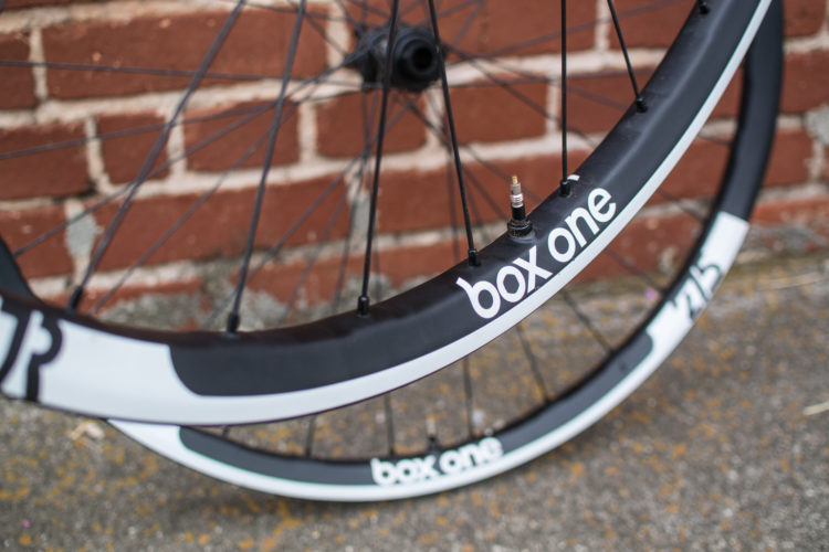 Close-up of a bicycle wheel with a black rim featuring the brand name "box one" in white lettering. The wheel is partially in focus, with a textured brick wall in the background. The valve for inflation is visible on the rim.