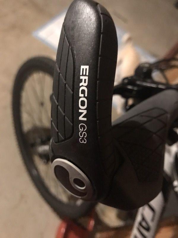 Cannondale Catalyst 2: Close-up image of an ergonomic bicycle grip labeled "ERGON GS3," featuring a textured surface for better grip, mounted on a bicycle handlebar. The background is slightly blurred, showing parts of a bike and a garage setting.