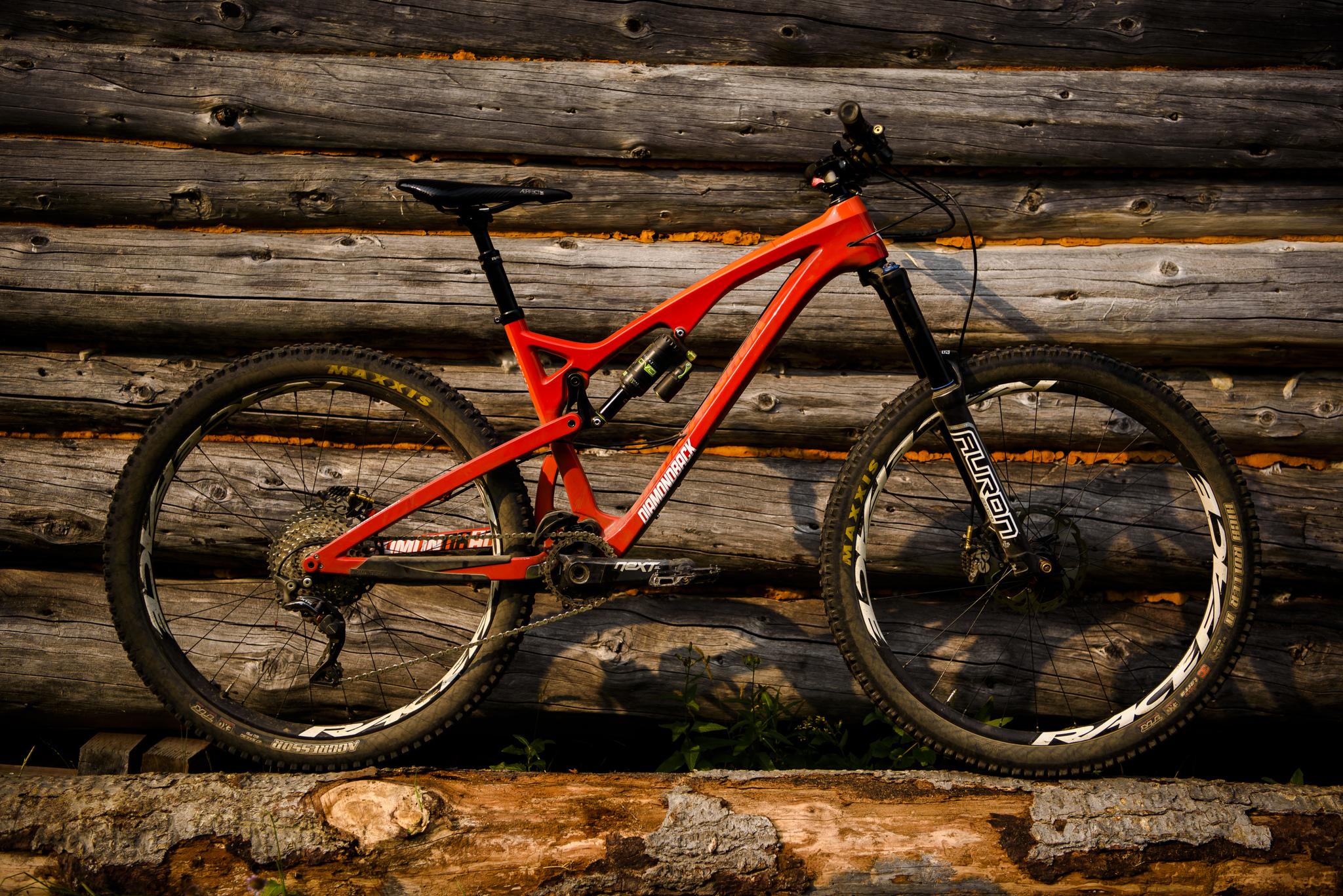 Diamondback Release Carbon: A vibrant red mountain bike positioned against a rustic wooden log wall. The bike features thick tires and suspension components, showcasing a sturdy design suitable for off-road terrain. The setting highlights a blend of nature and outdoor adventure.
