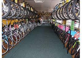 Interior of a bicycle shop featuring rows of bicycles displayed on both sides, with shelves holding bicycle wheels above. The shop has a green carpet and bright lighting.
