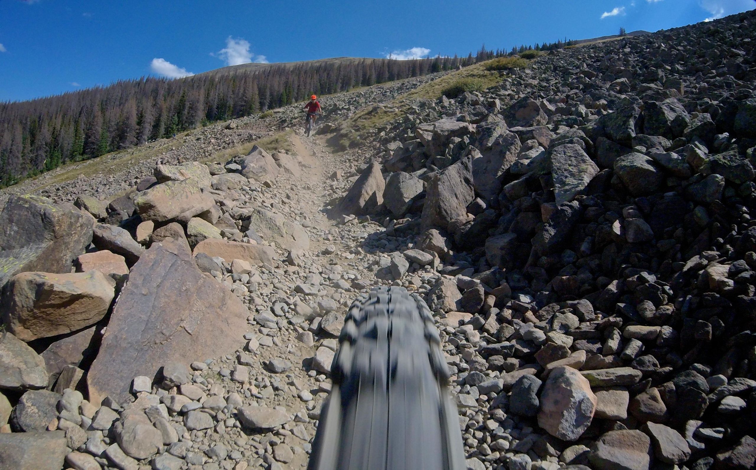 A close-up view of a mountain bike tire rolling over a rocky trail, with a rider in the background navigating the uneven terrain. The scene is set against a backdrop of blue skies and distant mountains, illustrating an adventurous outdoor environment. Monarch Crest Trail mountain bike trail.