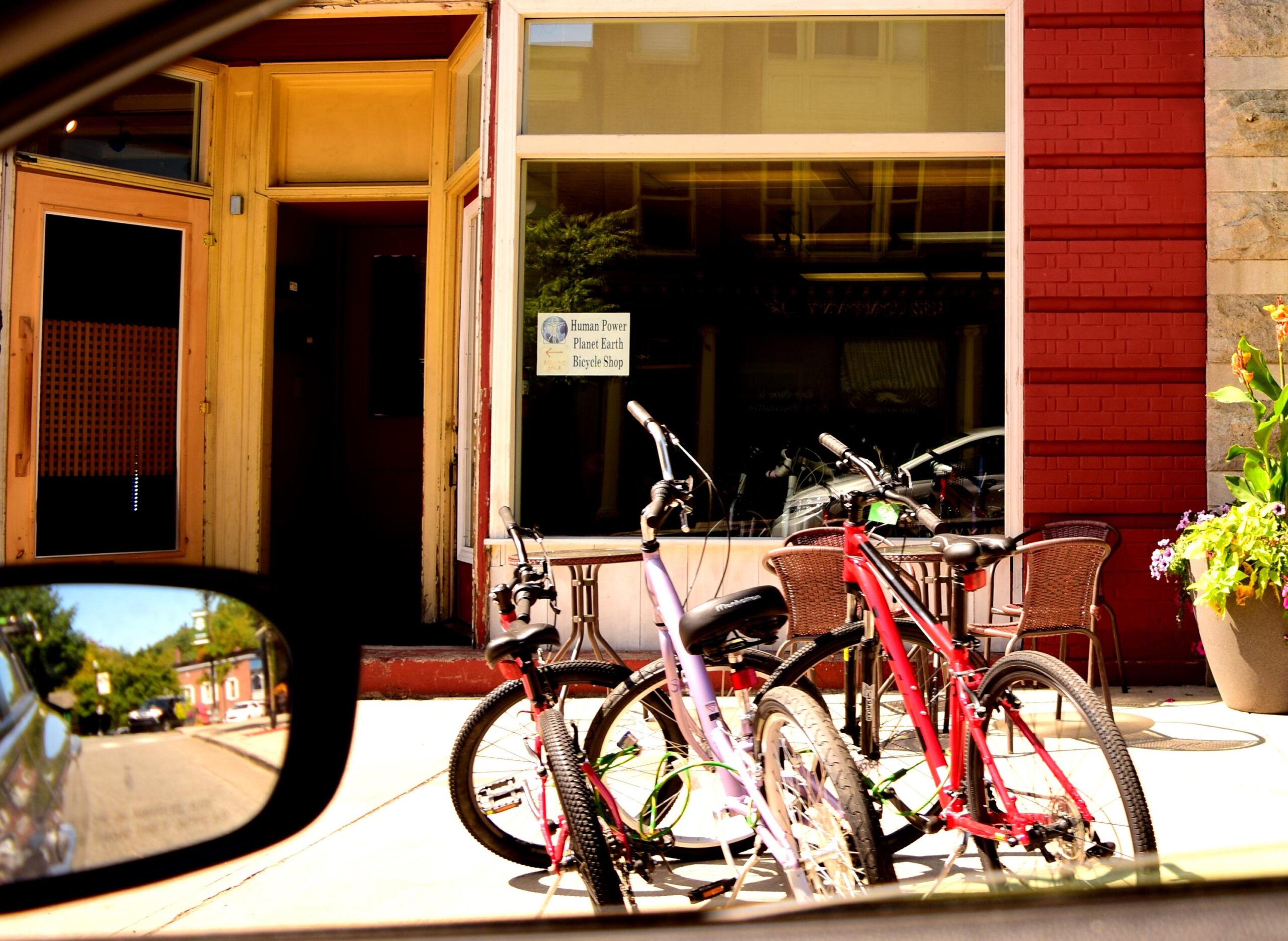 Bicycles parked in front of a bicycle shop with a sign that reads "Human Power Planet Earth Bicycle Shop." The scene includes outdoor seating and a planter with flowers, viewed from inside a vehicle.