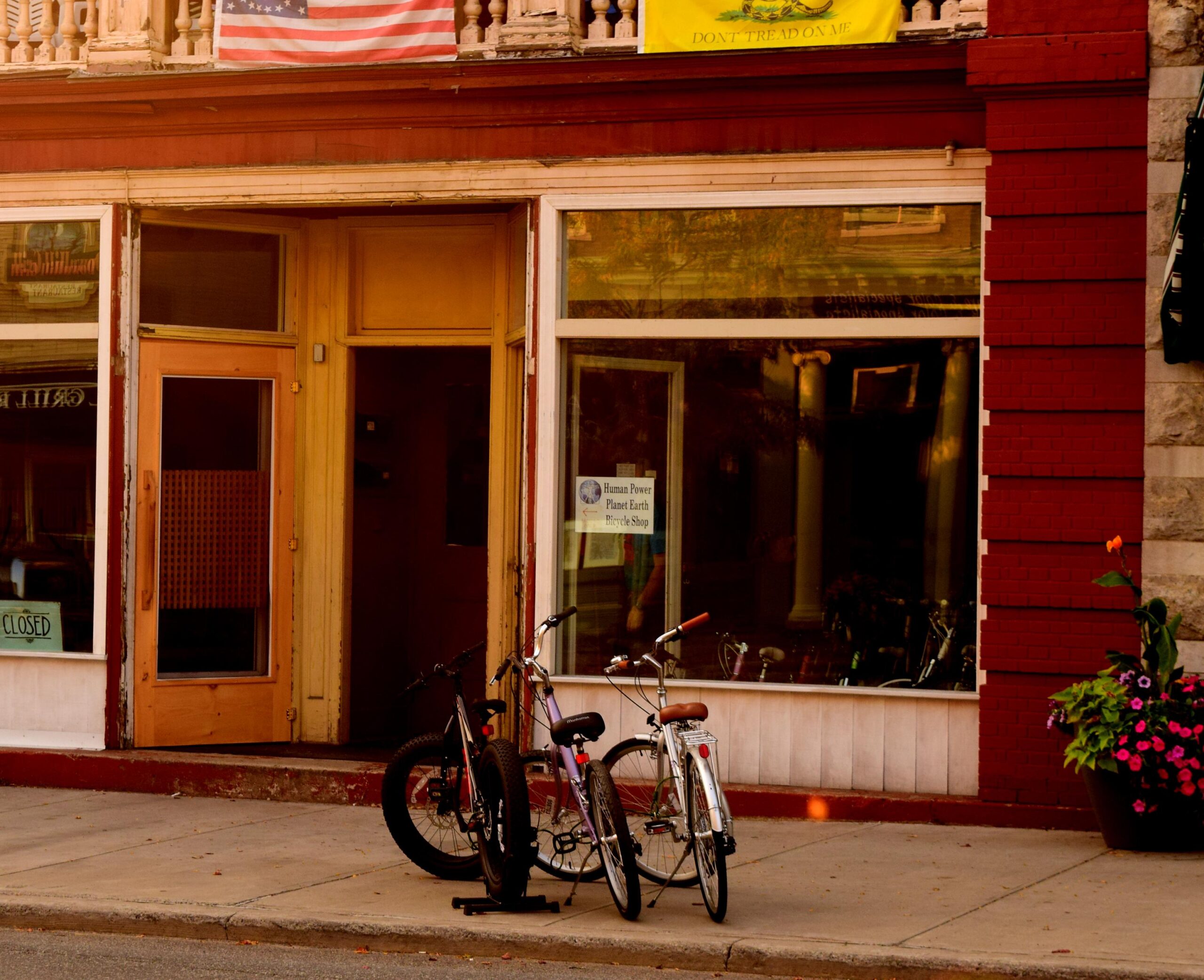 A storefront featuring large windows, with a sign indicating that the shop is closed. Two bicycles are parked outside on the sidewalk. Above the entrance, there is an American flag and a "Don