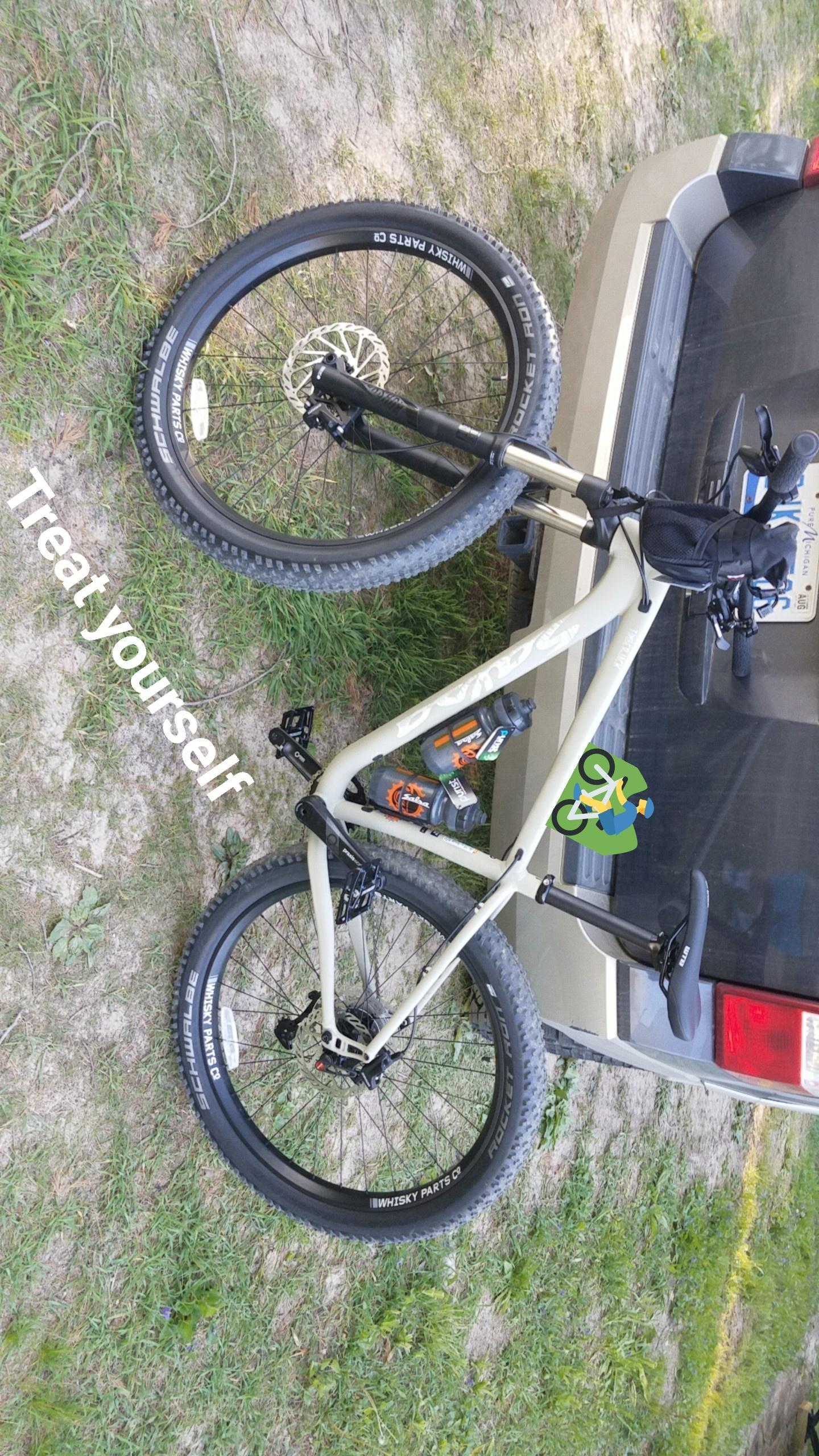 Salsa Timberjack: A mountain bike with black tires and a light-colored frame is parked on a grassy area next to a vehicle's rear. A water bottle is mounted on the bike, and the text "Treat yourself" is displayed alongside it. The setting appears outdoors, with a natural background.