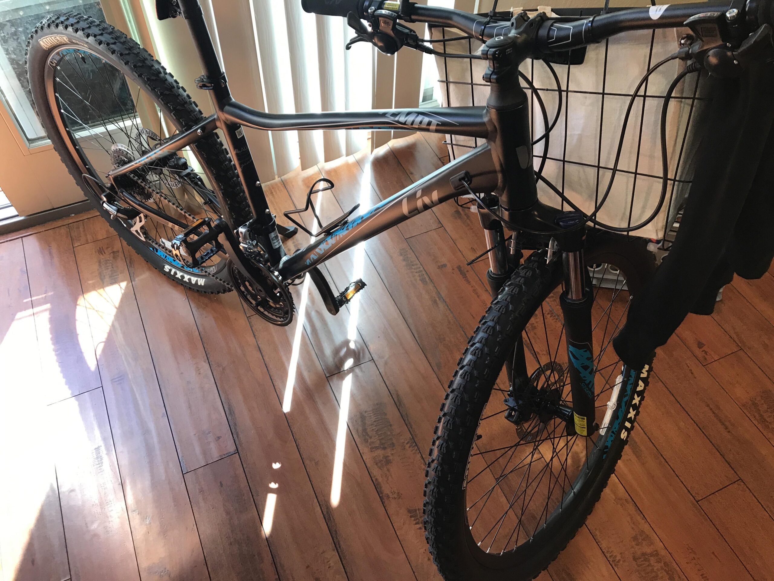 LIV LIV Tempt 3 2018: Image of a mountain bike leaning against a wall indoors. The bike features thick, treaded tires and a dark frame with blue accents. Sunlight casts diagonal shadows across the wooden floor, and a basket is attached to the handlebars.