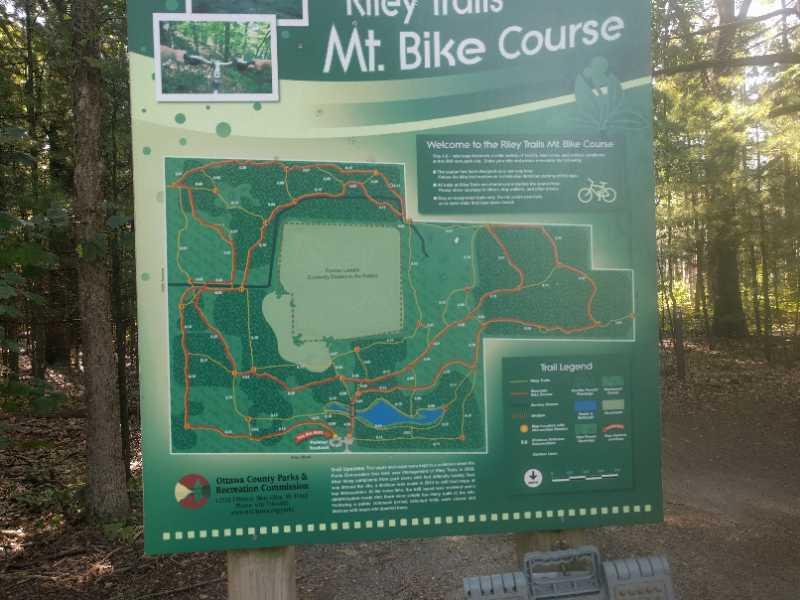 Alt text: A sign displaying a map and information about the Riley Trails Mountain Bike Course, featuring a trail layout with various paths marked in red and different terrain types indicated. The sign includes a welcome message and guidelines for using the bike course, set against a wooded background. Riley Trails mountain bike trail.