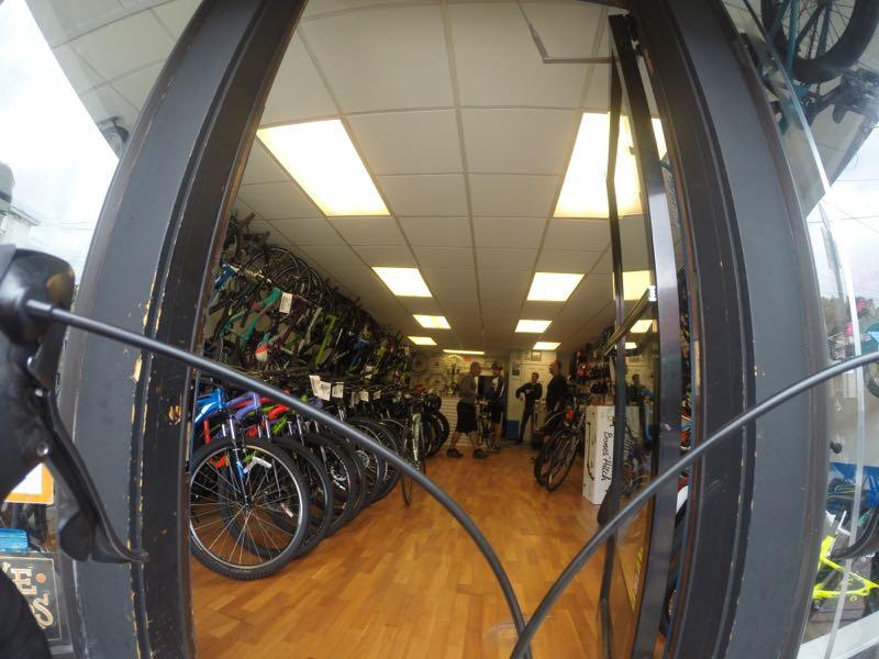 A view from outside a bicycle shop, showcasing a variety of bicycles displayed inside. The interior features wooden flooring, bright overhead lighting, and staff interacting with customers. Bicycles of different colors and styles are lined along the walls and on the floor.