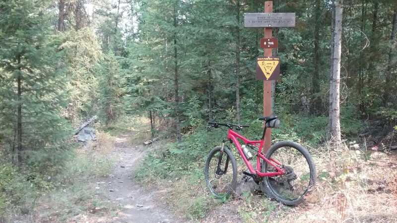 A red mountain bike rests against a post marking a hiking or biking trail in a wooded area. The sign includes a warning symbol and information about the trail, surrounded by tall trees and underbrush. The dirt path leads deeper into the forest. Clark Fork River Trail #223 mountain bike trail.