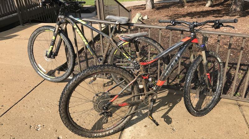 Two mountain bikes are leaning against a metal railing, both showing signs of mud and dirt on their frames and tires, indicating recent use on rugged trails. The bike on the right features a predominantly black and red frame, while the one on the left has a black and yellow design. The background includes a landscaped area with trees and a pathway. Addison Oaks mountain bike trail.