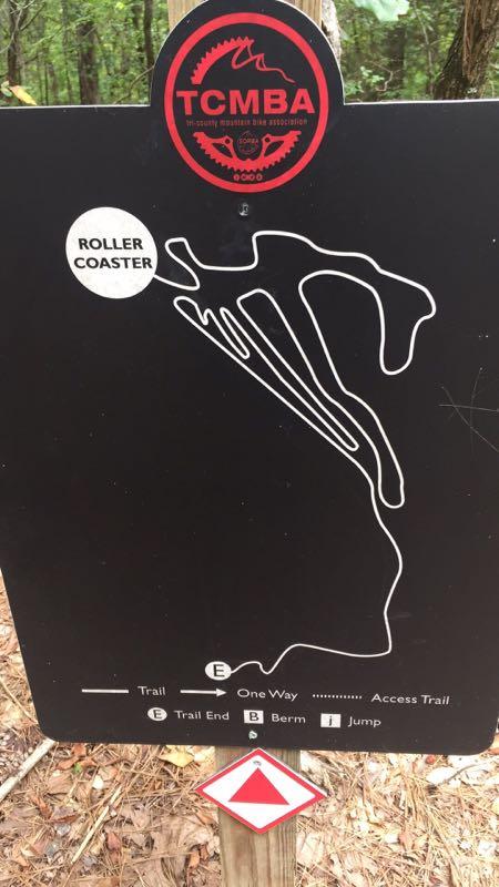 Image of a trail sign for a mountain biking route labeled "Roller Coaster." The sign features a black background with a white graphic depicting the trail's layout. It includes symbols indicating trail directions and features such as "One Way," "Berm," "Jump," and "Trail End." A logo from the "TCMBA" (Tri-County Mountain Bike Association) is displayed at the top. The setting appears to be surrounded by trees, indicating a natural outdoor environment. The Ridgeland Trails mountain bike trail.