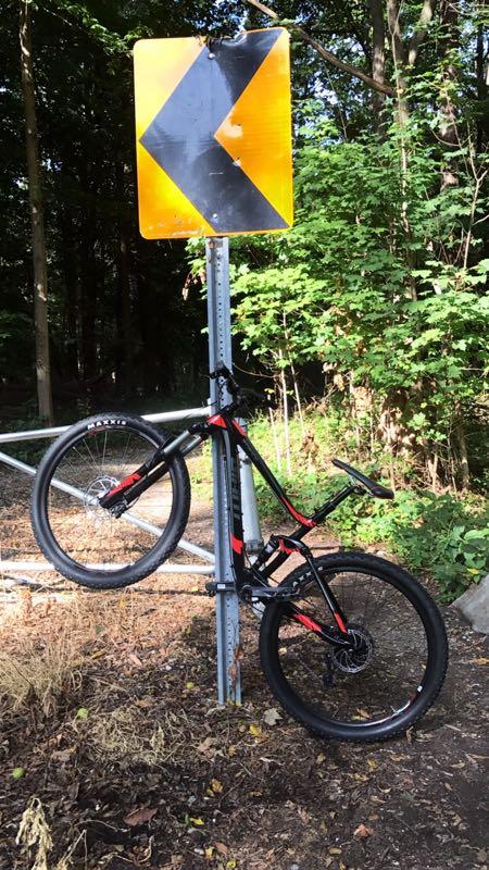 A black and red mountain bike leaning against a caution sign indicating a sharp turn, surrounded by greenery and a dirt path. Novi Tree Farm (Lakeshore Park) mountain bike trail.