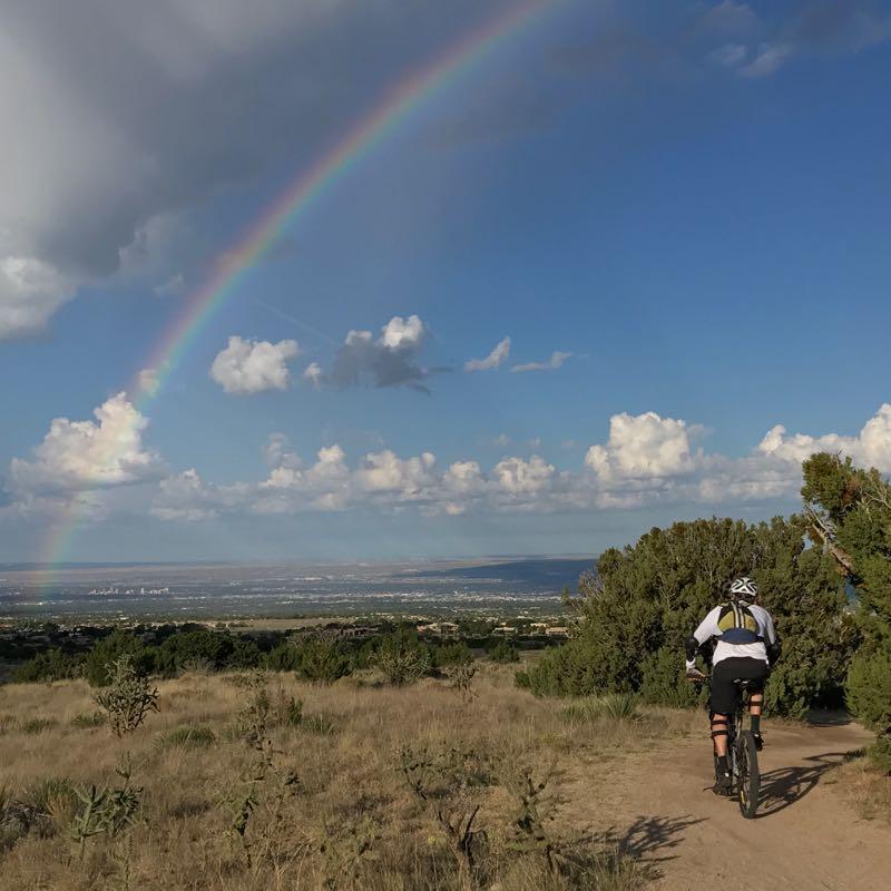 A mountain biker rides along a dirt trail overlooking a panoramic view of a valley. A rainbow arcs across a blue sky filled with fluffy white clouds, casting a vibrant backdrop to the scene. Green shrubs and dry grass line the trail, enhancing the natural beauty of the landscape. Elena Gallegos Open Space / North Foothills mountain bike trail.