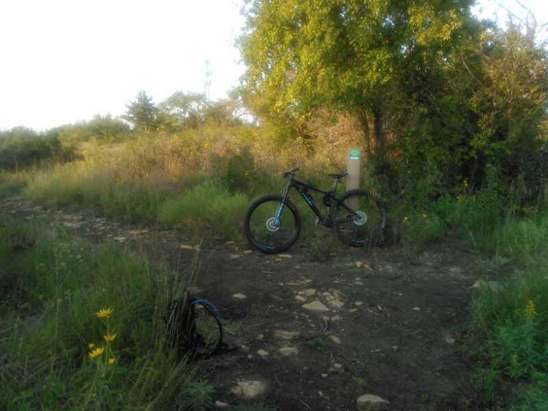 A mountain bike resting on a dirt trail surrounded by tall grass and vegetation, with a trail marker visible in the background. Skyline Park - Burnetts Mound Trail mountain bike trail.