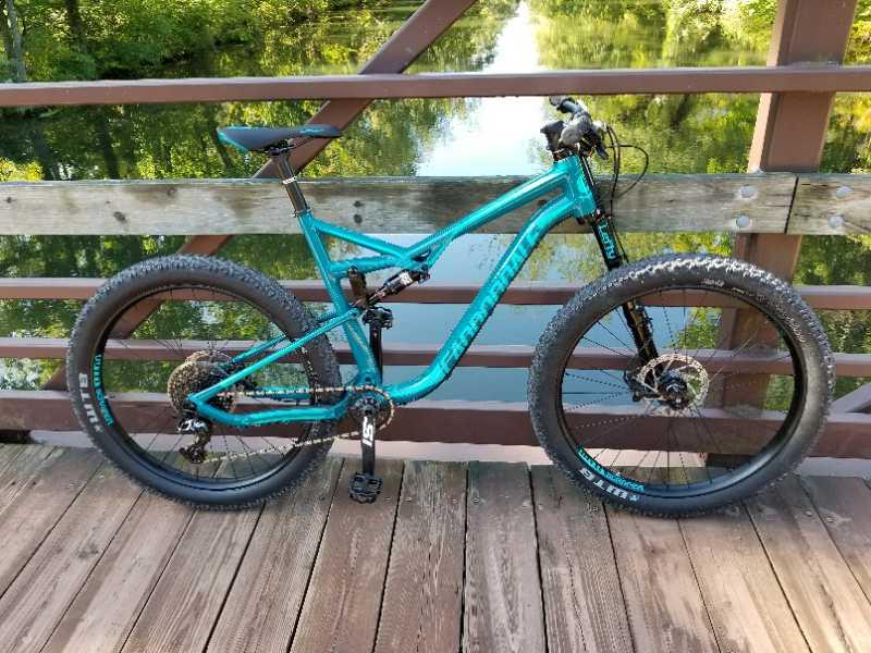 A blue mountain bike is parked on a wooden bridge with a reflective water surface in the background, surrounded by lush green trees. The bike features thick tires and a modern design, showcasing its suitability for off-road terrain. Six Mile Run mountain bike trail.