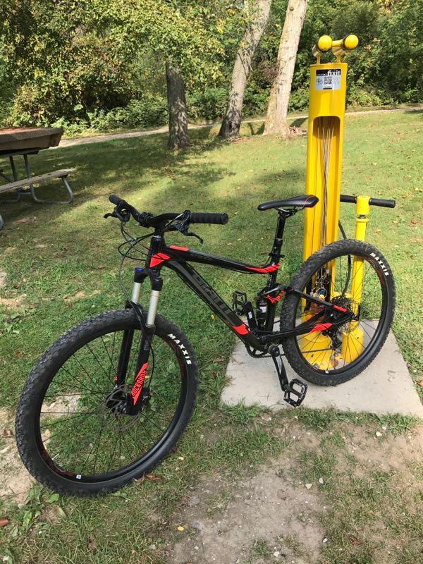 A black and red mountain bike is parked next to a yellow bike repair station in a grassy area with trees in the background. The bike is equipped with wider tires suitable for off-road riding. Holdridge Recreation Area mountain bike trail.